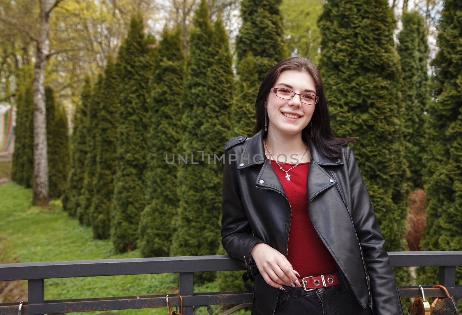 young beautiful brunette girl in black jacket and jeans stands on the embankment near river in city park on sunny spring day