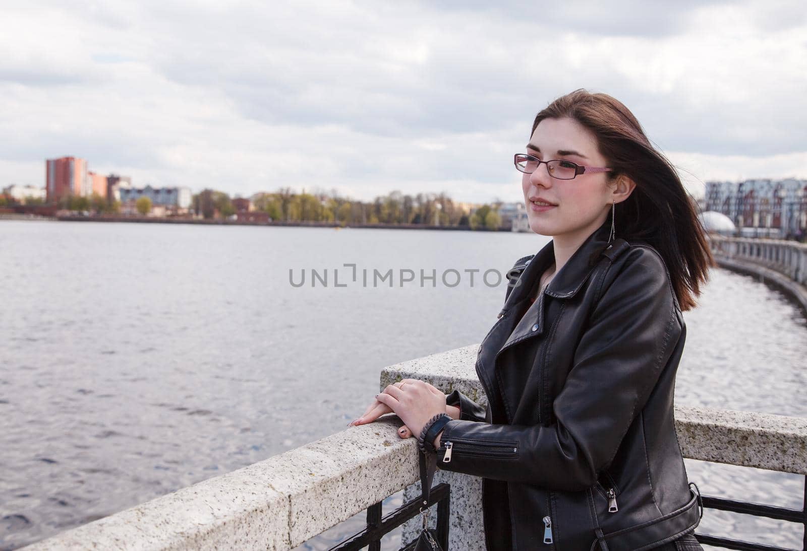 young beautiful brunette girl in black jacket and jeans stands on the embankment near lake in city park on sunny spring day