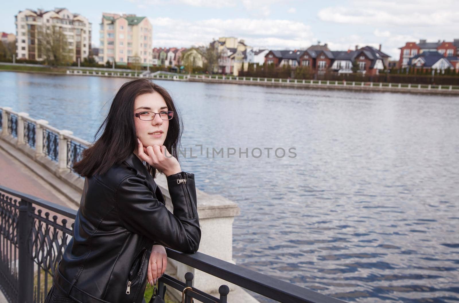 young beautiful brunette girl in black jacket and jeans stands on the embankment near lake in city park on sunny spring day
