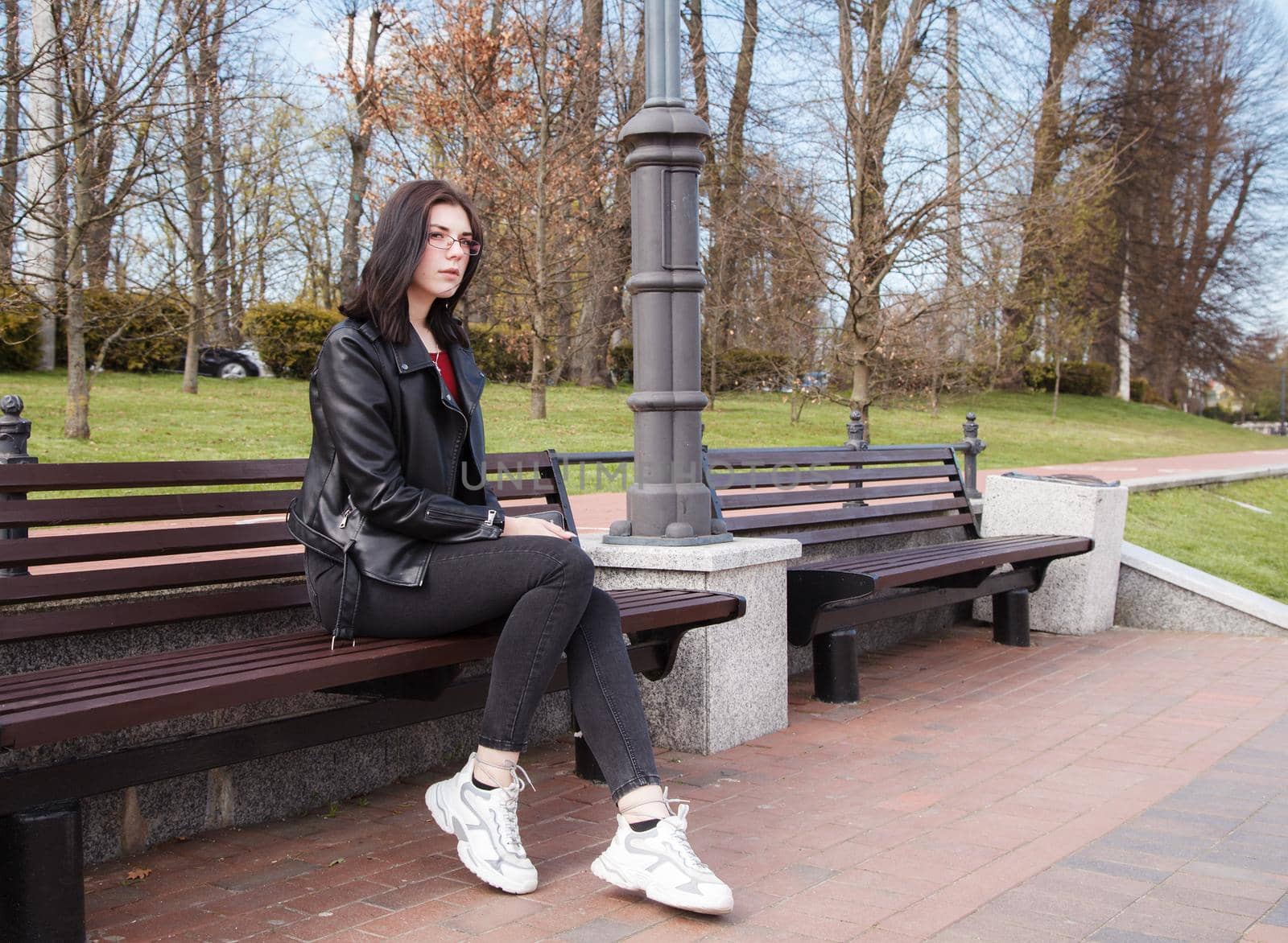 young girl in black jacket and jeans sits on a bench in a city park on sunny spring day