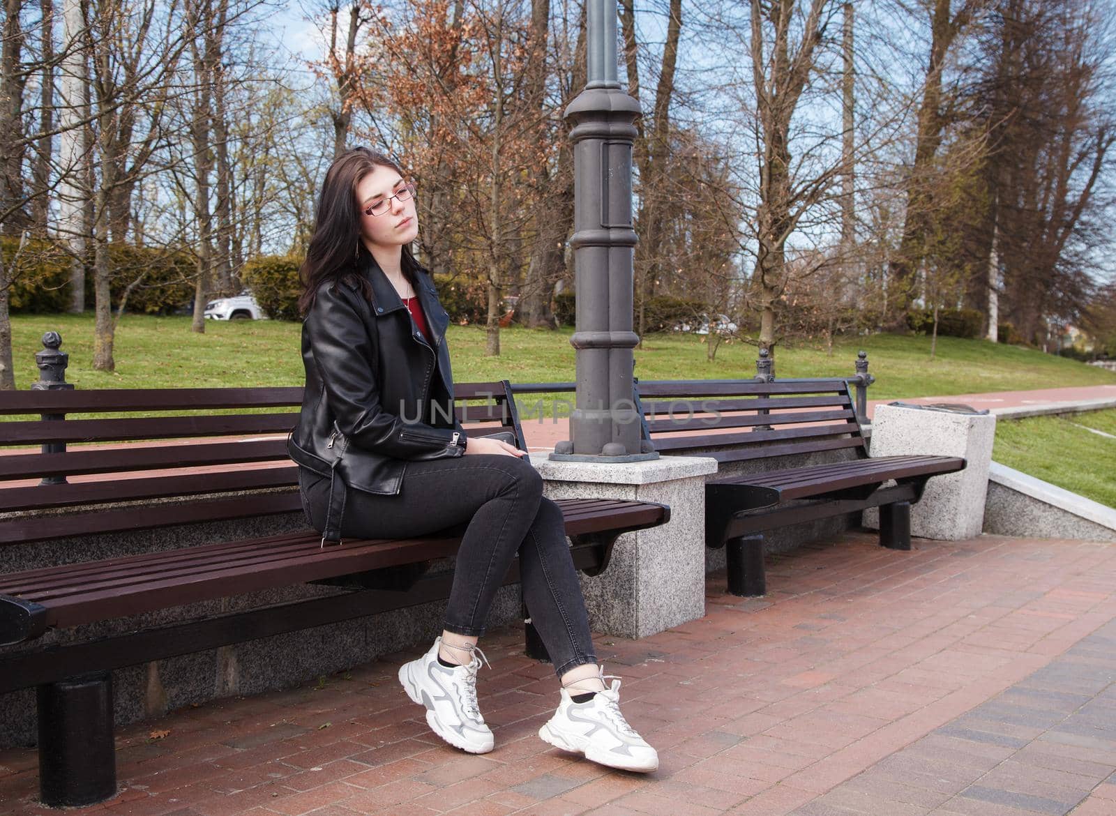 young girl in black jacket and jeans sits on a bench in a city park on sunny spring day