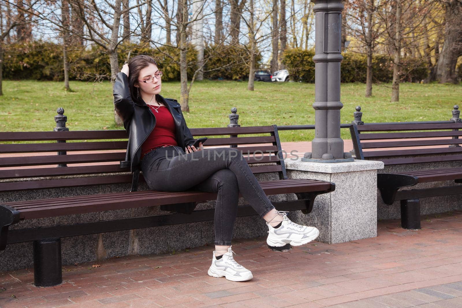 young girl in black jacket and jeans looking at smartphone display while sitting on bench in city park on sunny spring day