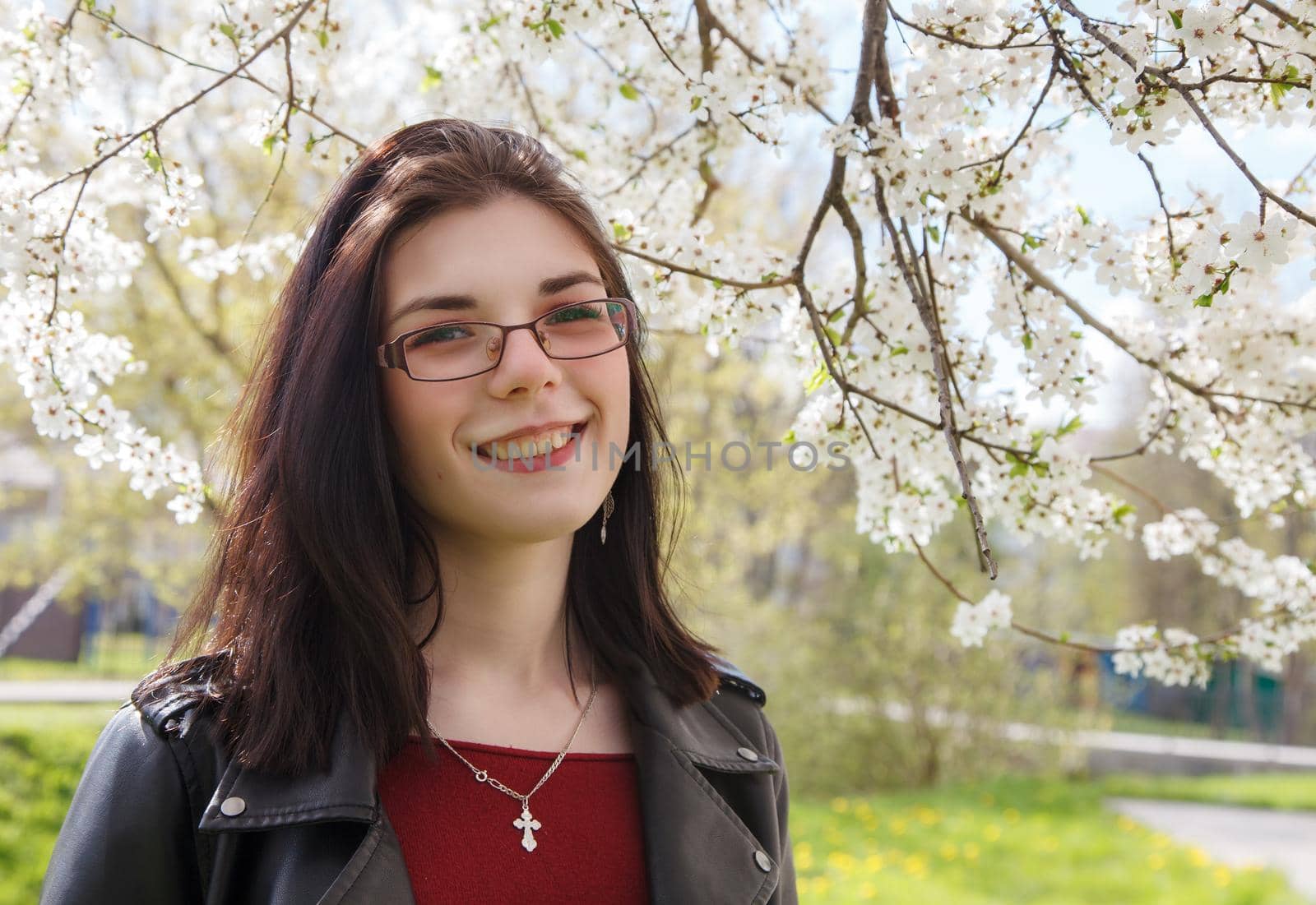 portrait of young beautiful brunette girl in black jacket and burgundy sweater standing near cherry blossoms on sunny spring day. outdoor closeup