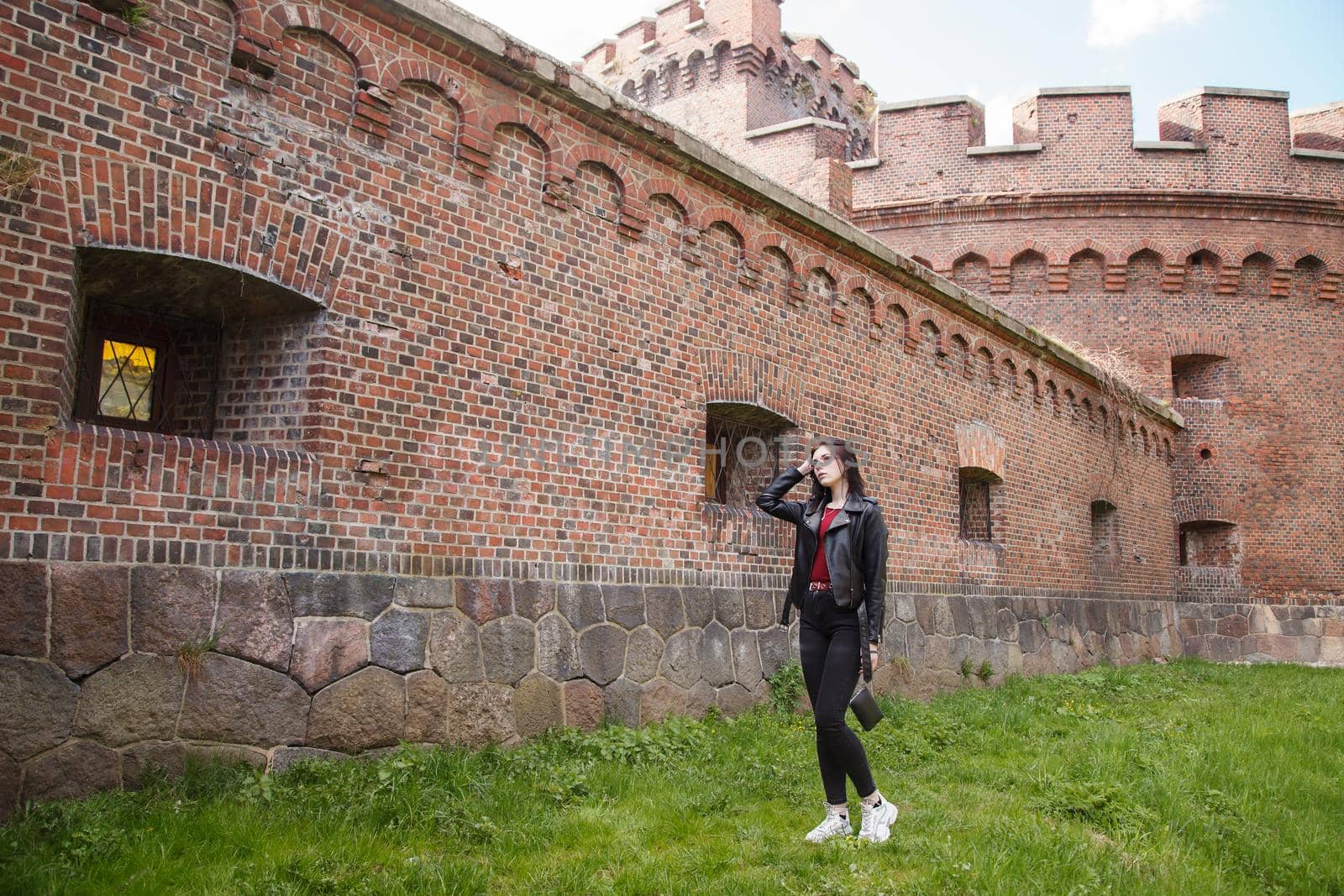 young beautiful girl in black jacket and jeans stands near the old fortress wall on sunny day