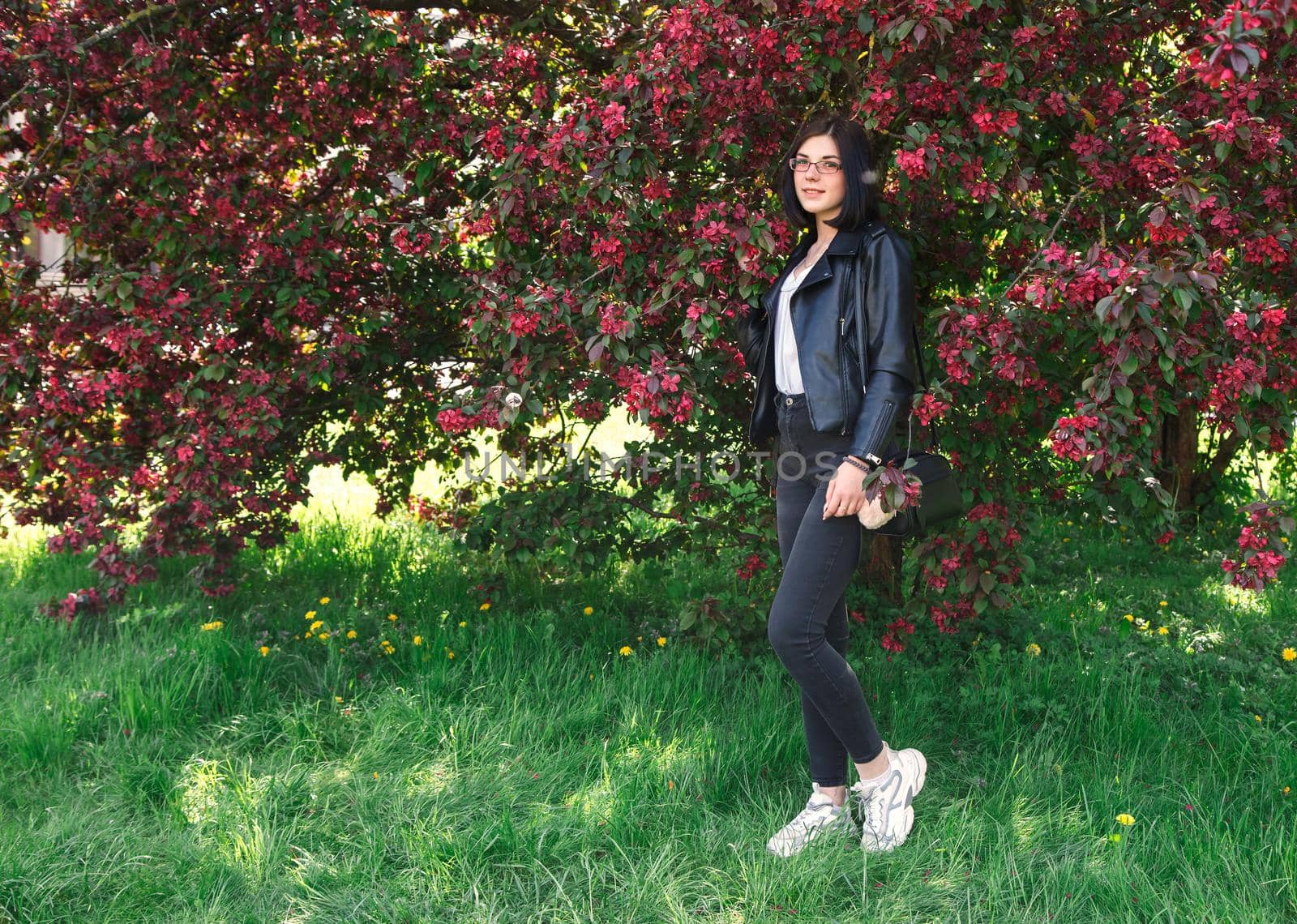 portrait of young beautiful brunette girl in black jacket standing near cherry blossoms on sunny spring day