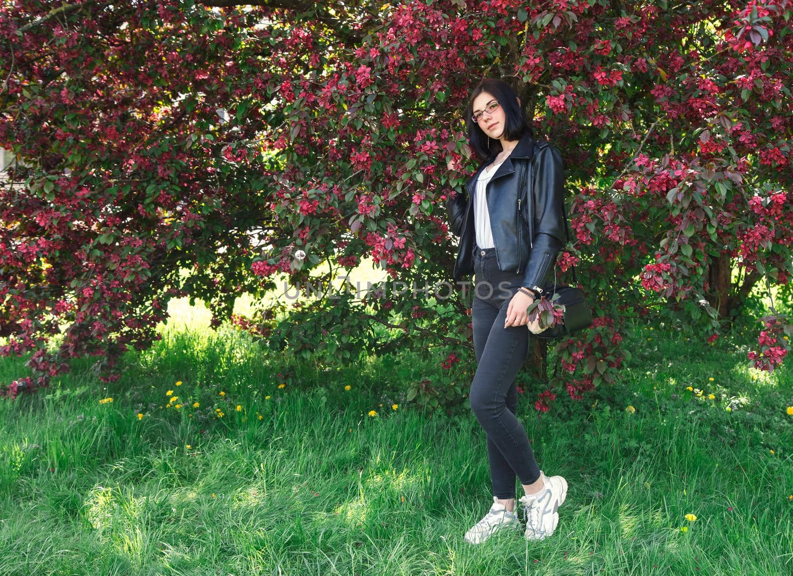 portrait of young beautiful brunette girl in black jacket standing near cherry blossoms on sunny spring day