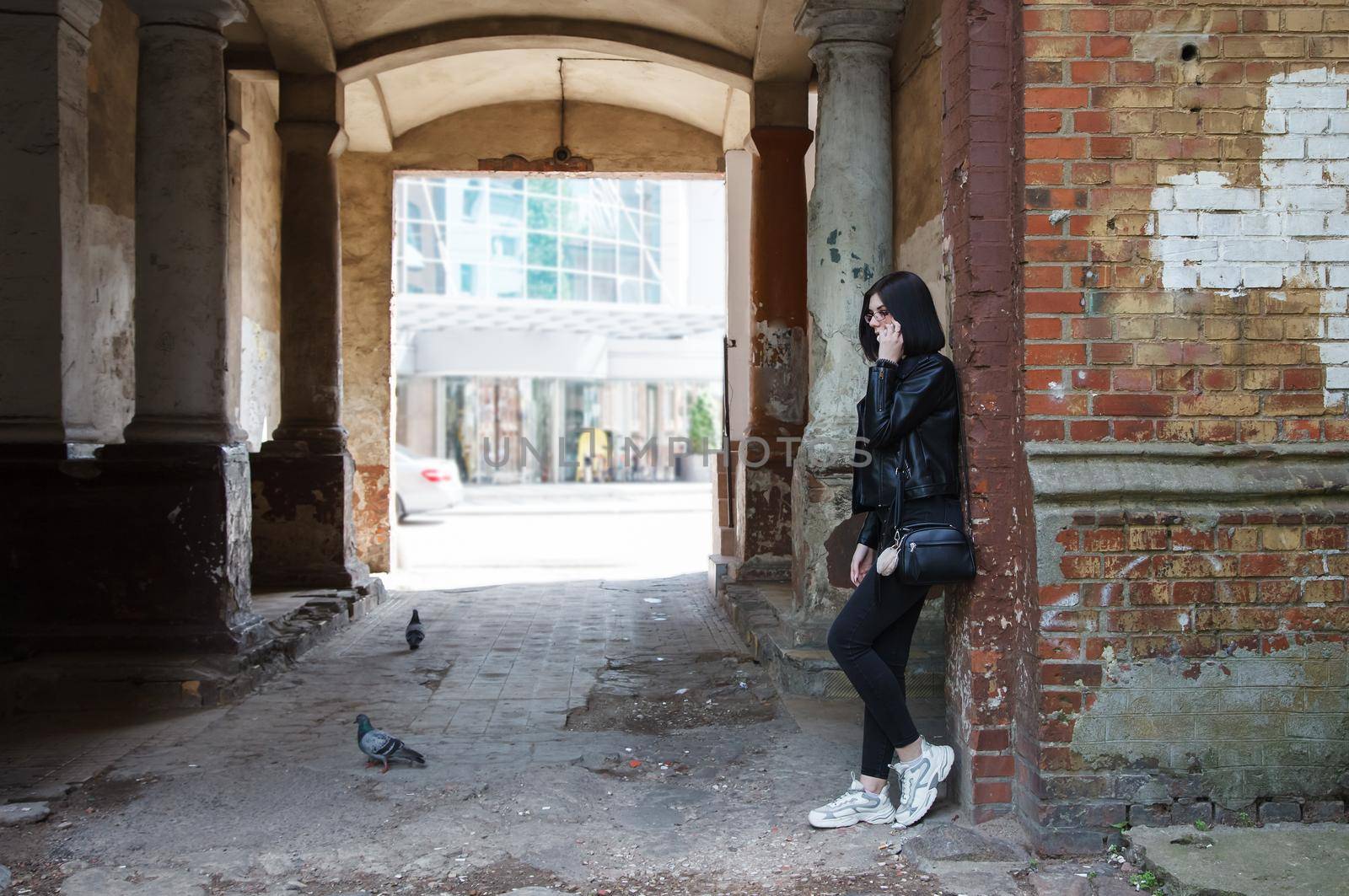 sad young girl in black jacket and jeans stands in old arch on city street on summer day