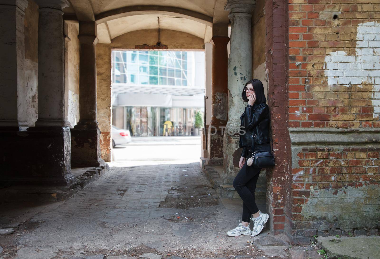 sad young girl in black jacket and jeans stands in old arch on city street on summer day