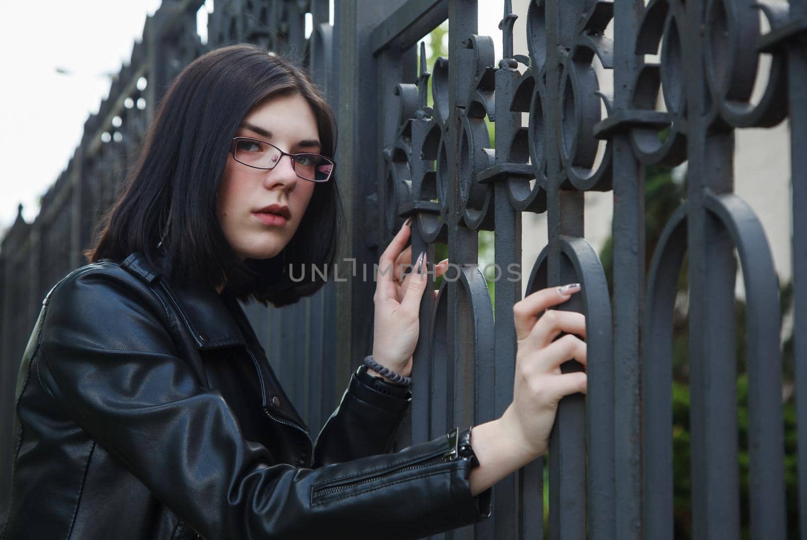 sad young girl in black jacket stands near metal fence on city street on summer day. closeup outdoor