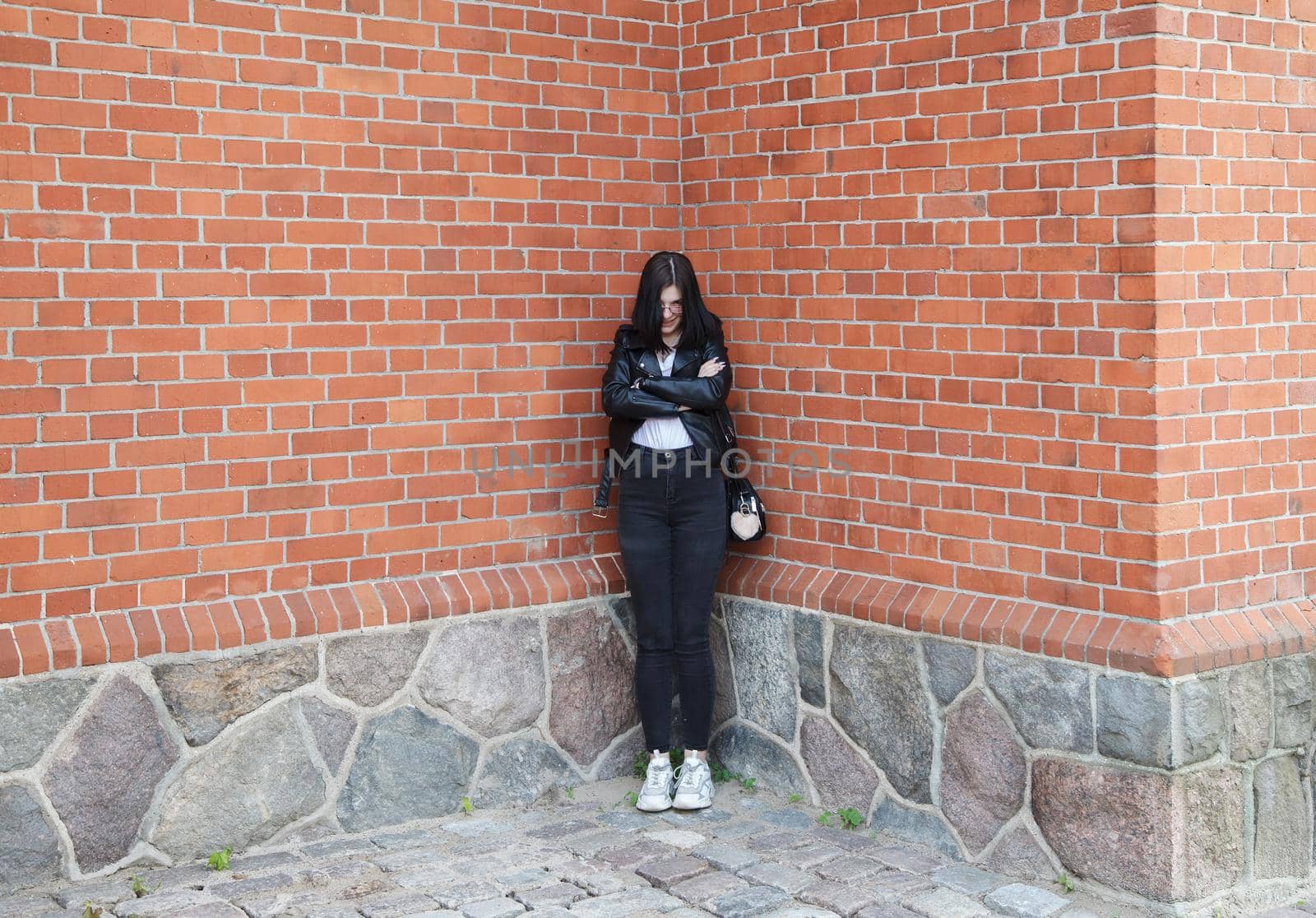 young beautiful brunette girl in black jacket and jeans stands near wall of gothic church on summer day