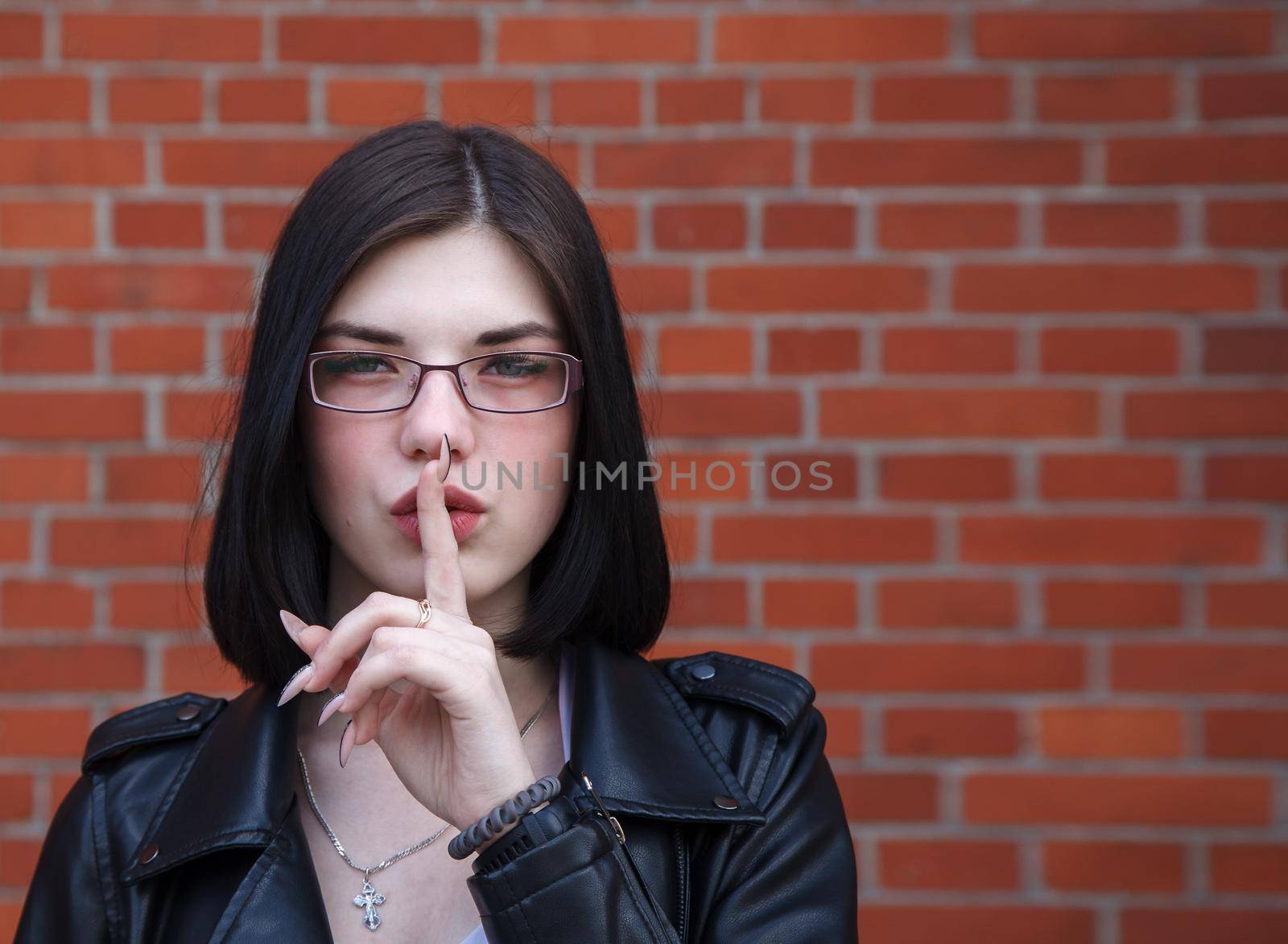 young beautiful brunette girl in black jacket shows a gesture quietly near brick wall. outdoor closeup