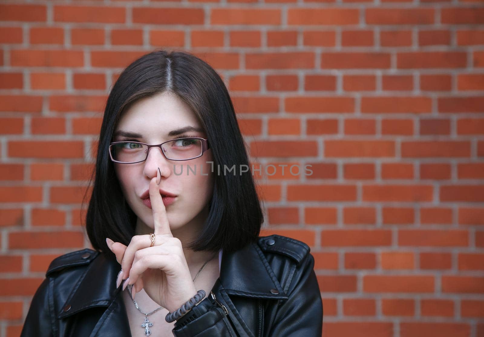 young beautiful brunette girl in black jacket shows a gesture quietly near brick wall. outdoor closeup