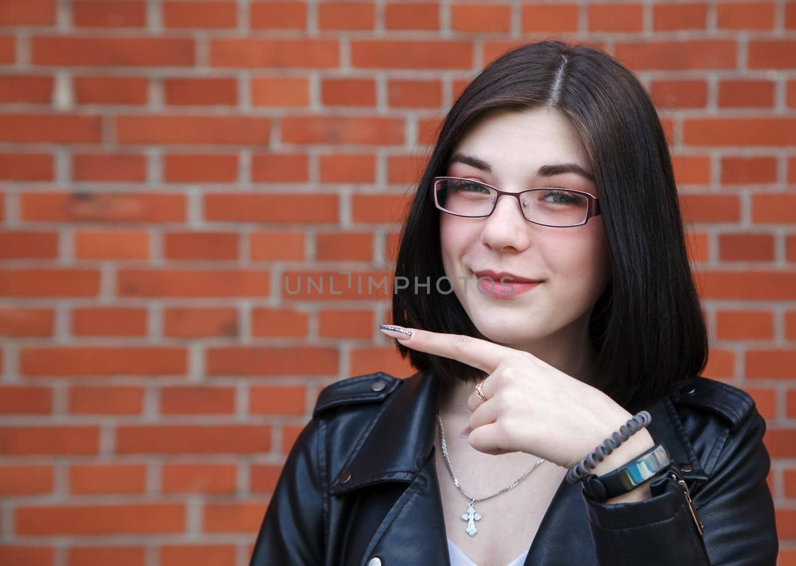 positive beautiful brunette girl in black jacket points to the side. presenting your product. outdoor closeup on brick wall background