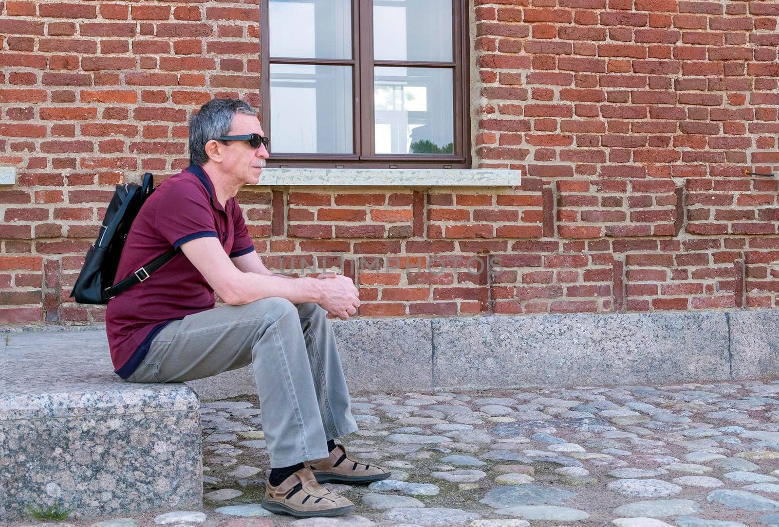 Adult pensioner with a backpack sits and rests against the backdrop of a red brick building by OlgaGubskaya