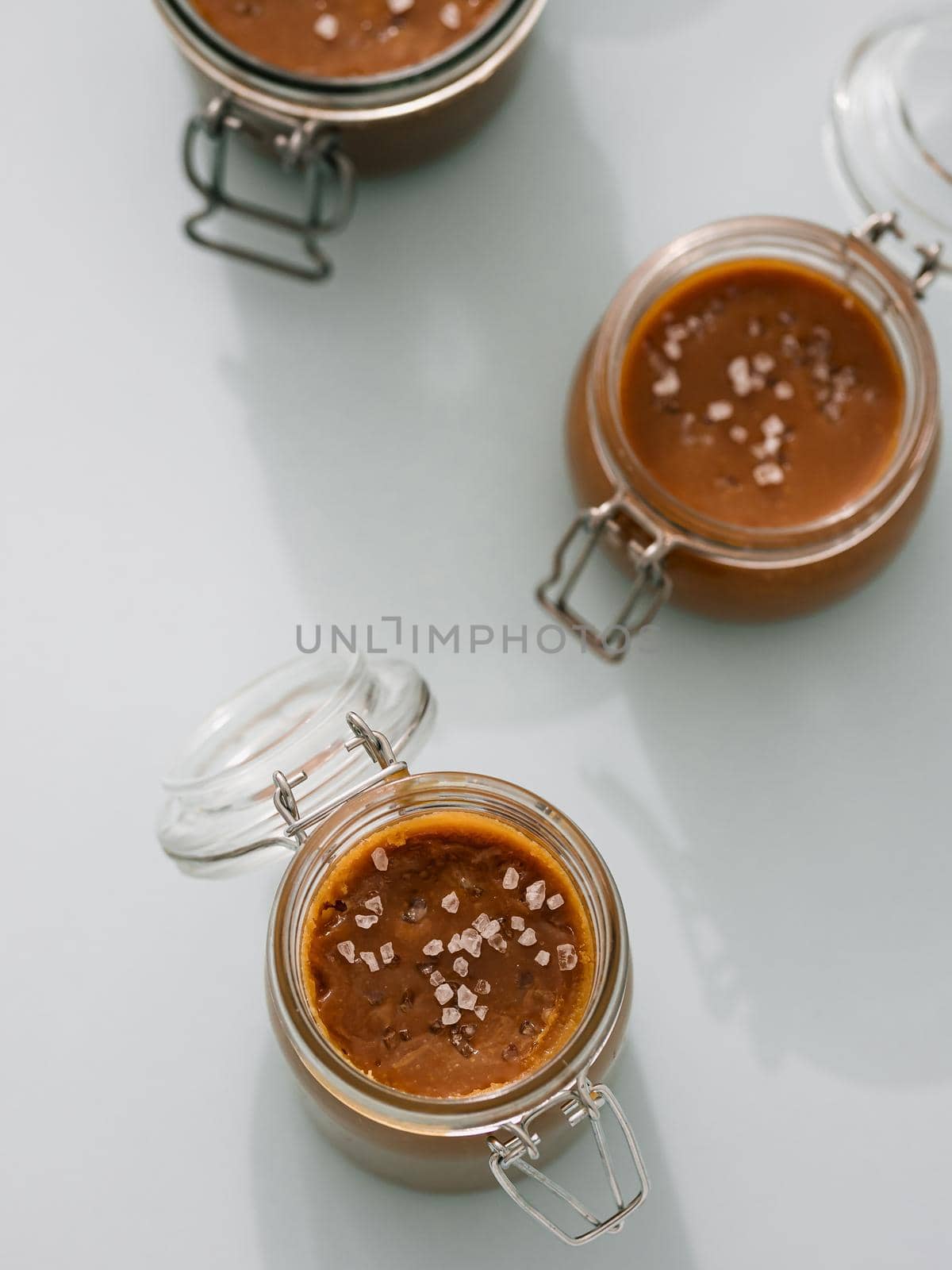 Salted caramel in glass jars, top view. Brown caramel or condensed milk with sea salt crystalls, shoot in bright or hard light. Light neutral background. Vertical