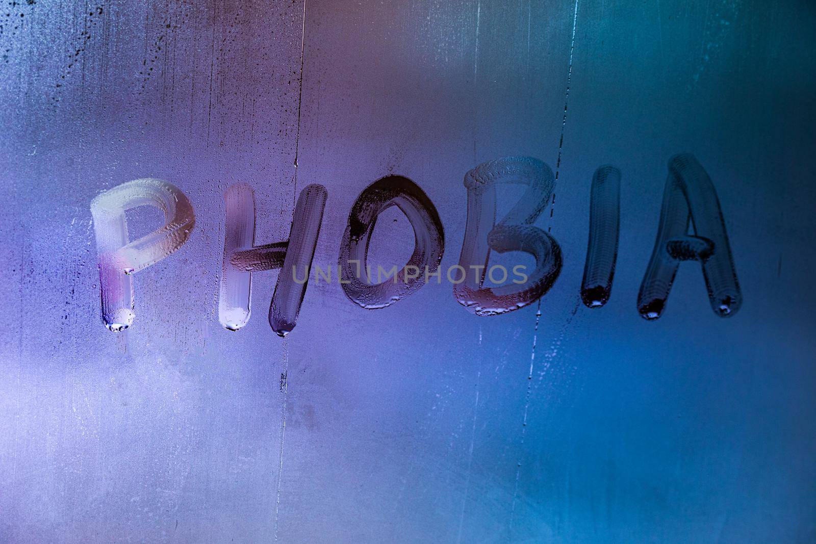 the word phobia handwritten on wet foggy window glass surface with blue background light.