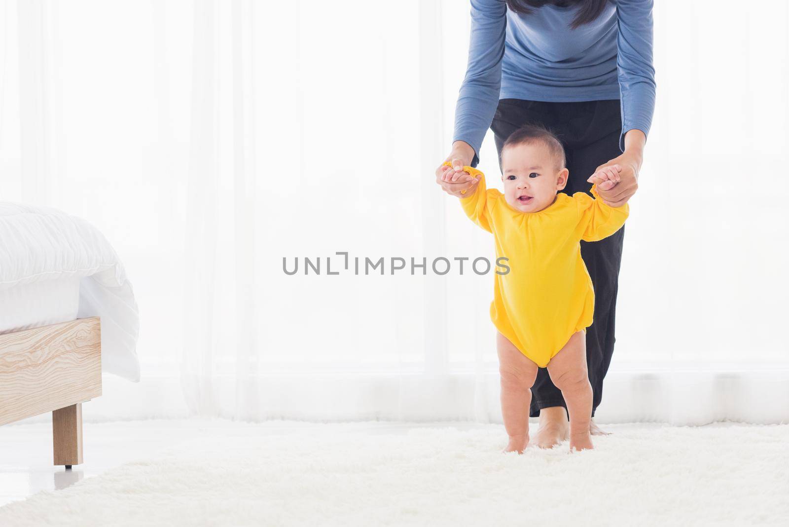 Asian little baby girl taking first steps learning to walk with mom help support the cute unstable walking toddler at home in bedroom. Happy family first steps parenthood concept