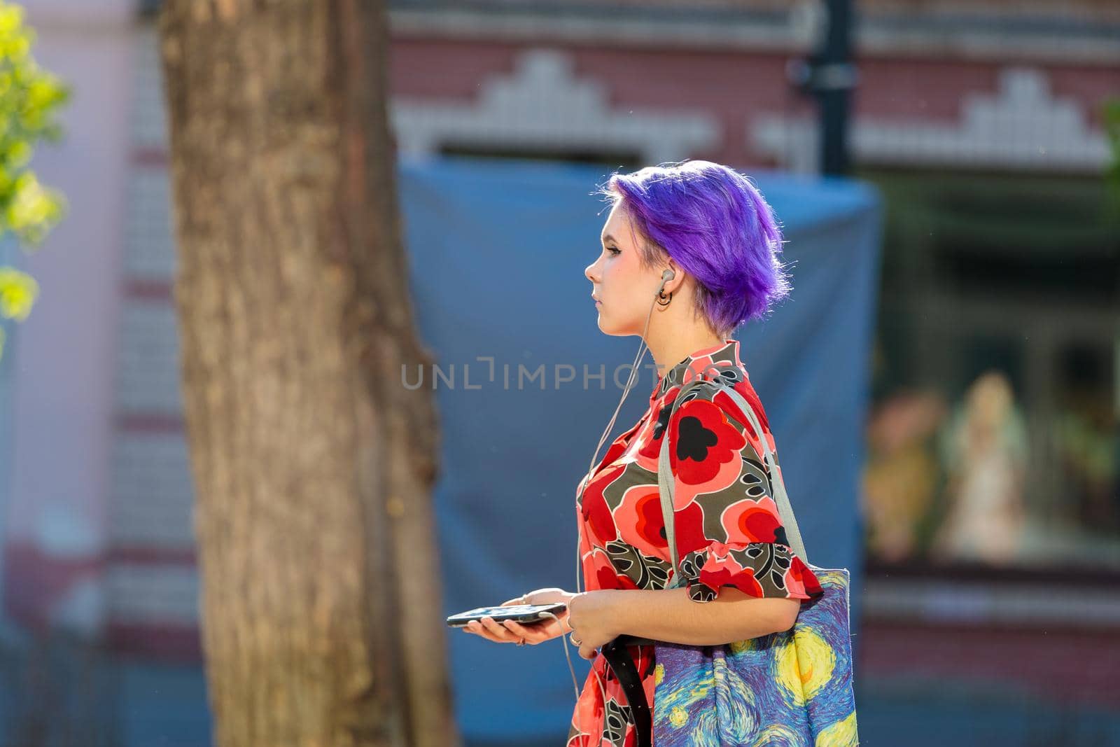 A girl walks along a city street with purple hair. Listens to music through headphones on the phone