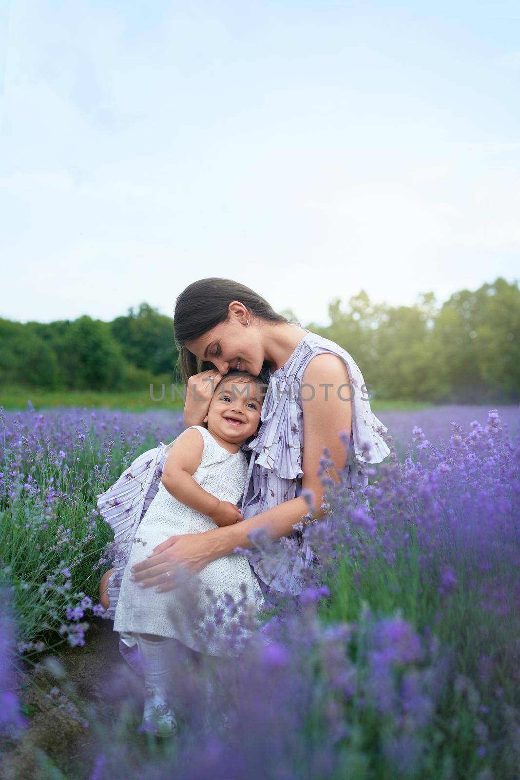 Side view of young mother wearing dress sitting between beautiful purple flowers. Woman posing with little smiling baby daughter on knees in lavender field. Concept of nature, motherhood.