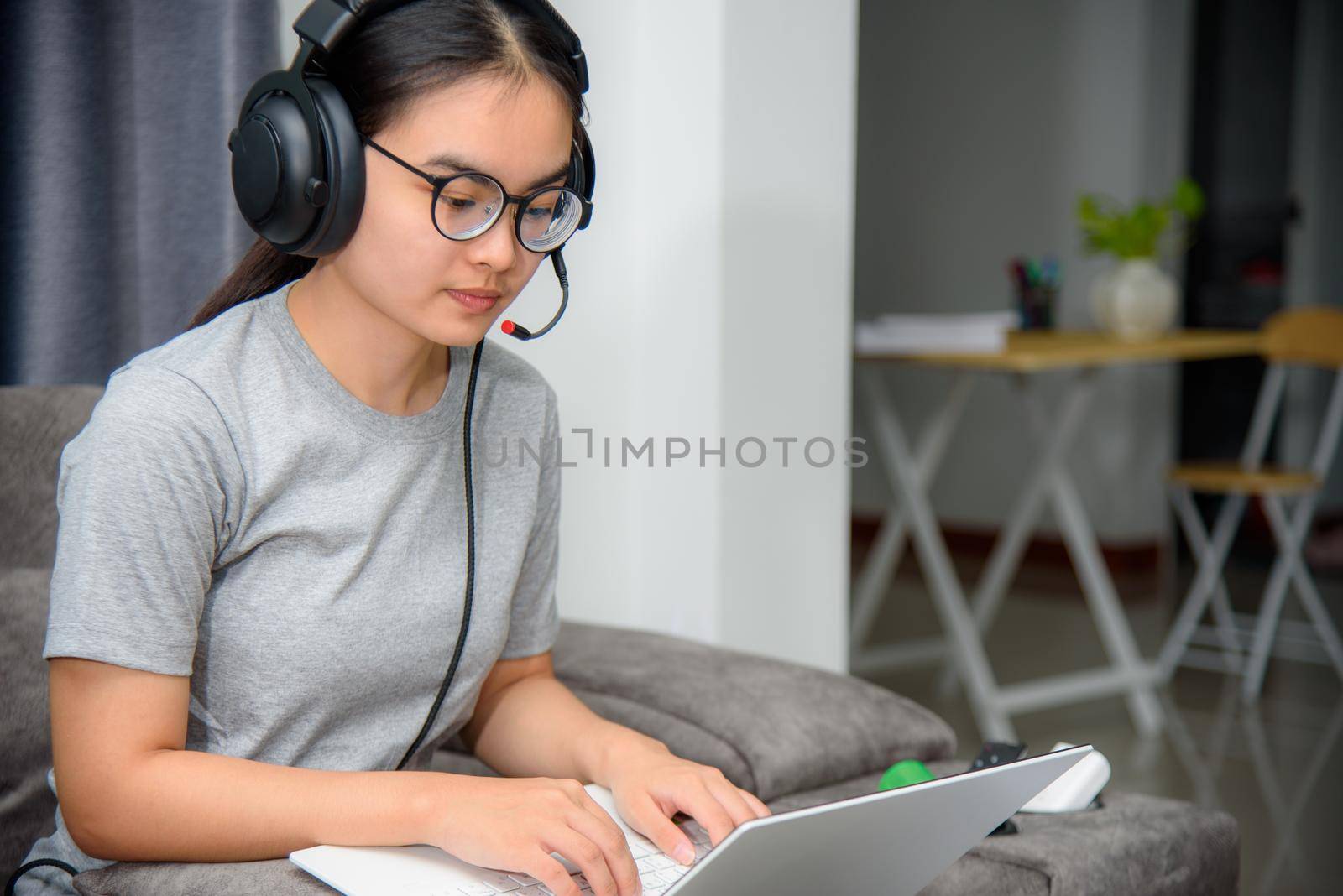 Asian young woman student with headphones sitting on the sofa look at laptop happy study online class college learning internet education, Teenage girl work distance on a laptop computer from home
