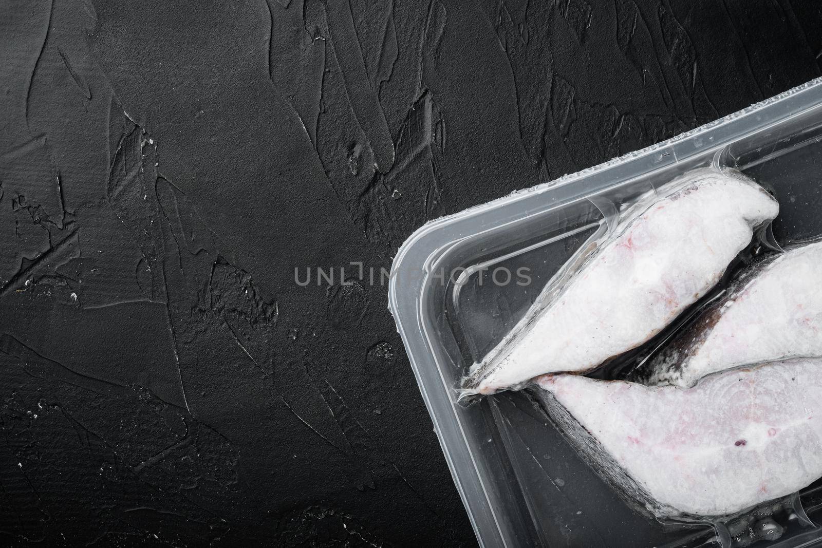 Frozen Greenland halibut steaks vacuum pack set, on black dark stone table background, top view flat lay, with copy space for text
