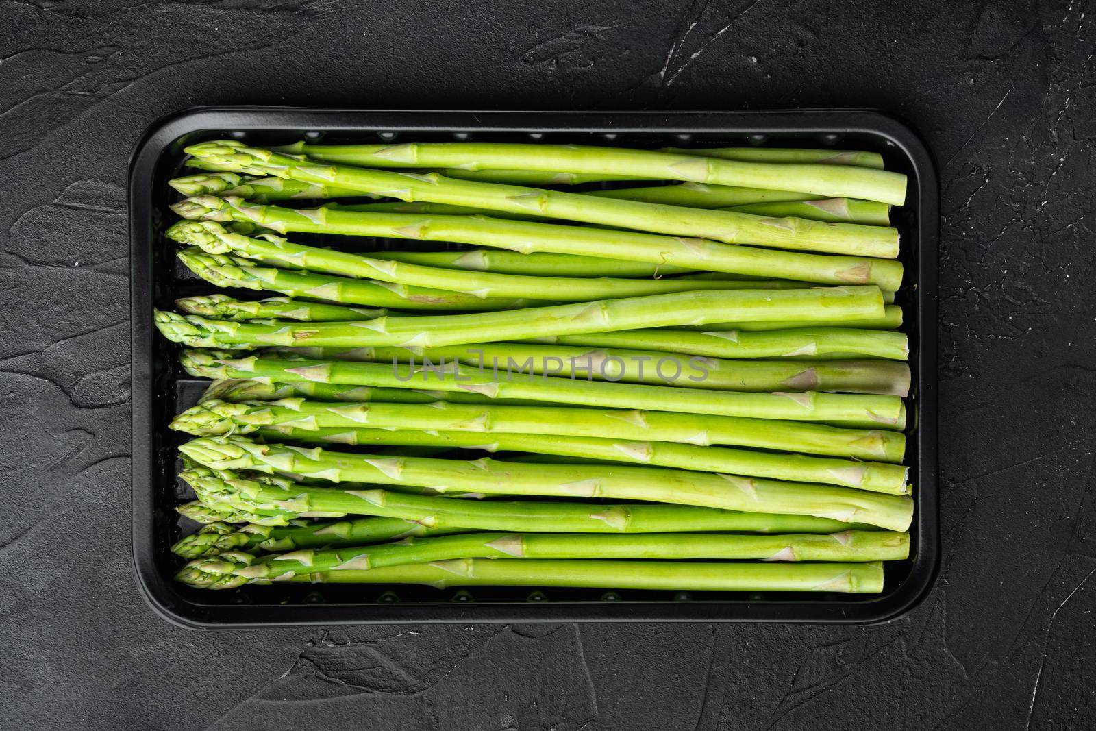 Bunches of fresh green asparagus set, in plastic market container, on black stone background, top view flat lay