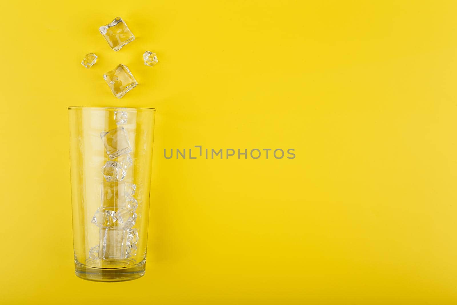 Creative flat lay with empty transparent glass with spilled ice cubes on yellow background with copy space. Concept of refreshing summer drinks and cocktails