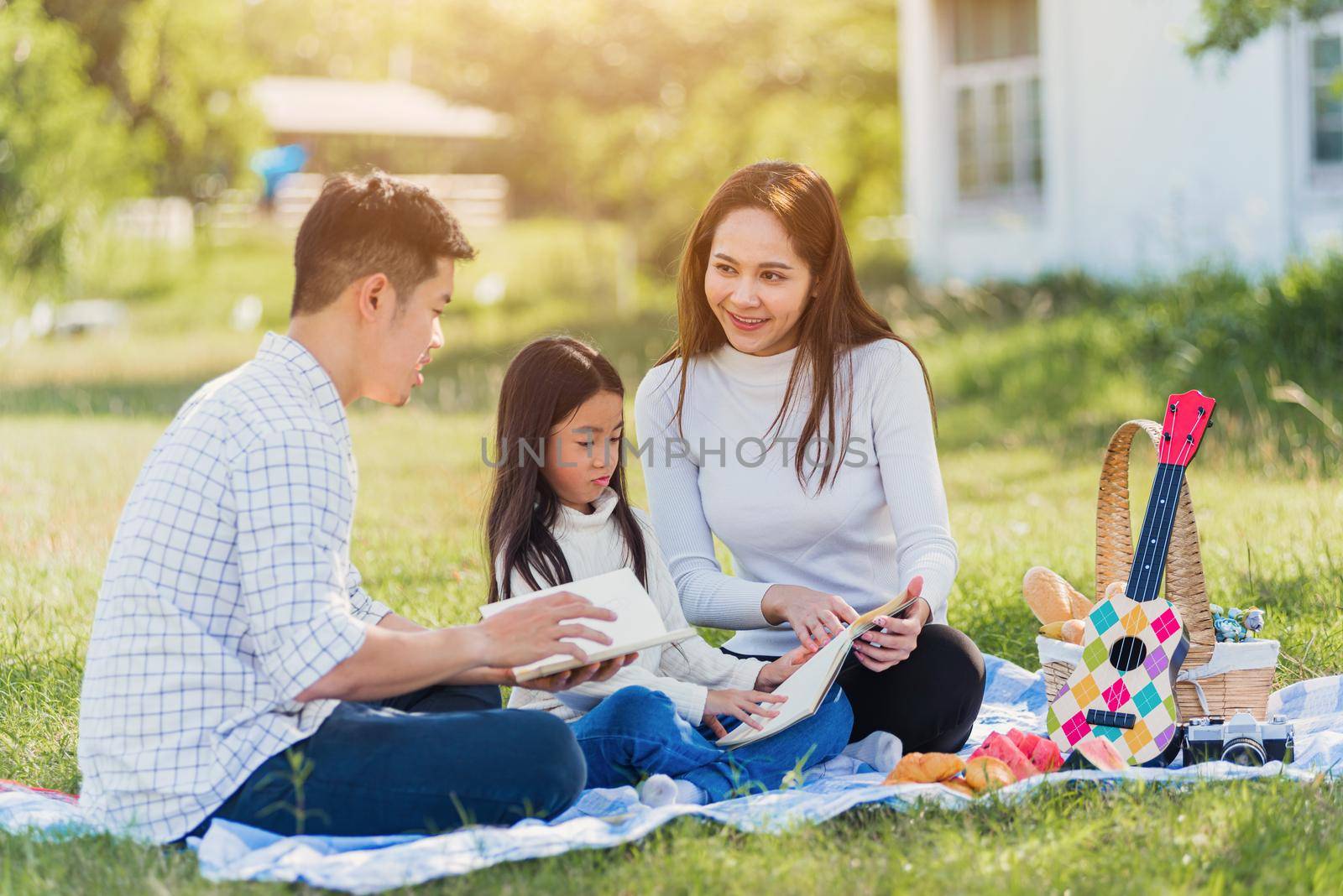 Asian family having fun and enjoying outdoor on picnic blanket reading book in park by Sorapop