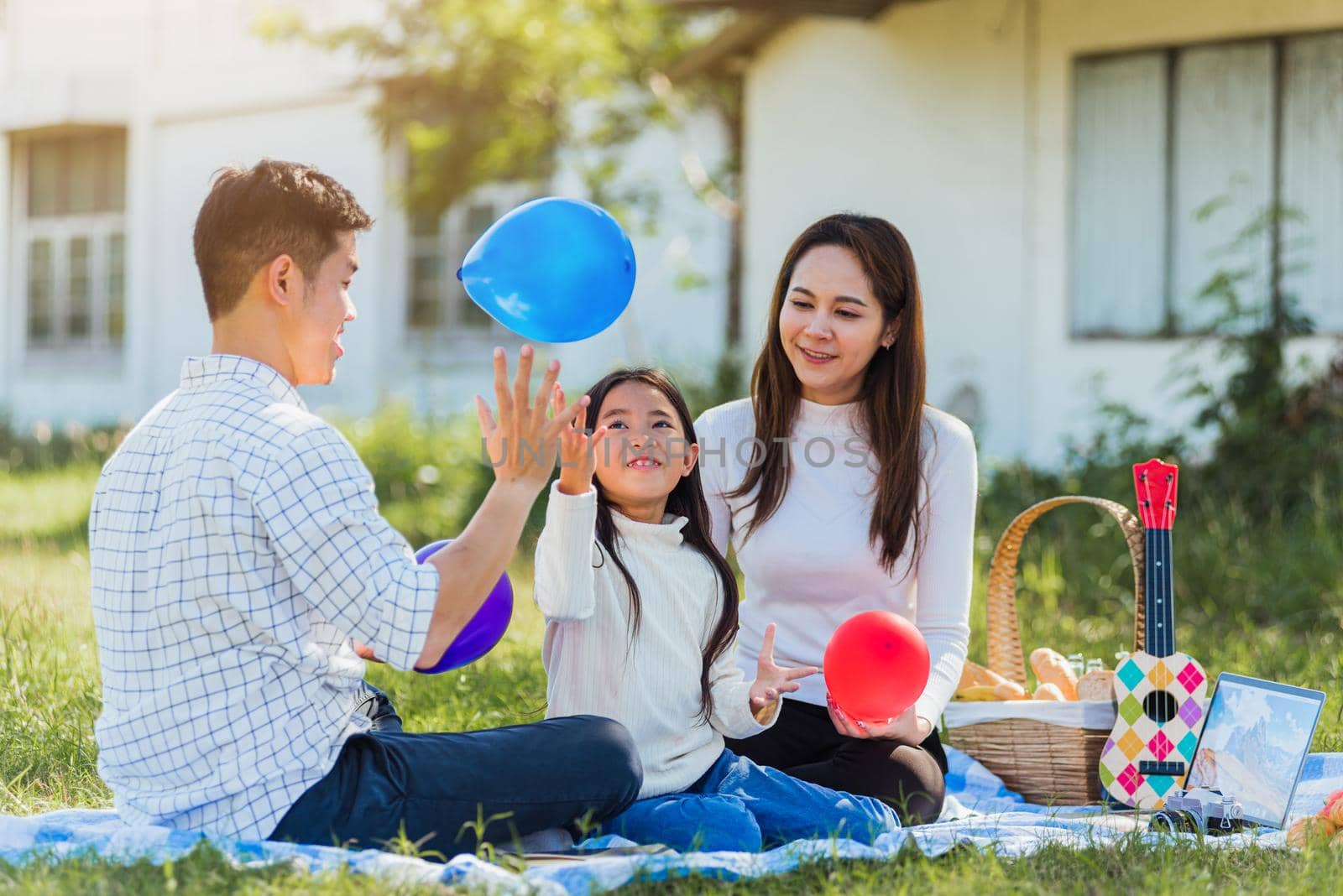 Happy Asian young family father, mother and child little girl having fun and enjoying outdoor sitting on picnic blanket playing balloons at summer garden spring park, Family relaxation concept