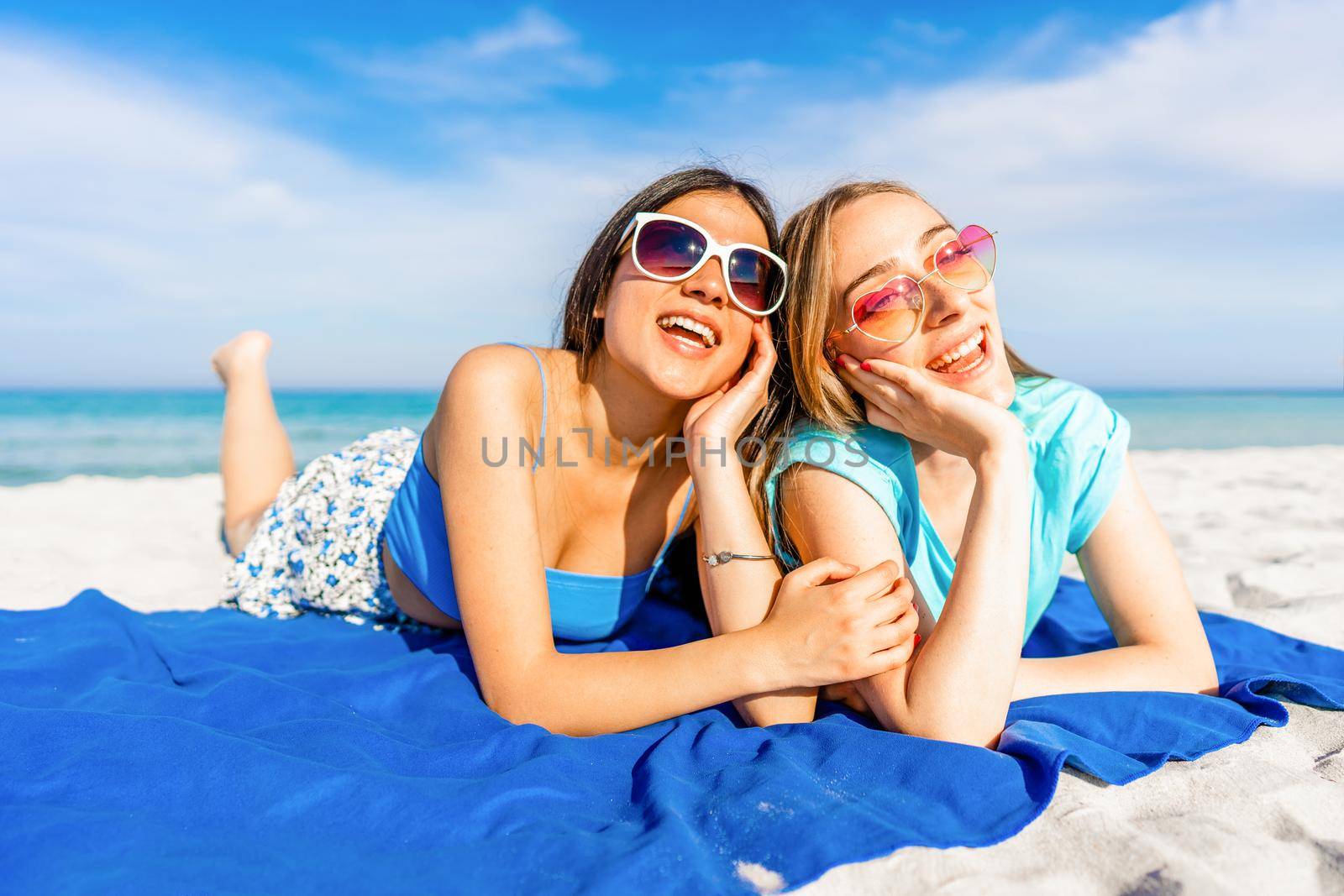 Front low view of Two happy beautiful girls lying on the tropical beach in summer vacation kissed by the sun with sunglasses smiling together. Blonde young woman with her best brunette girlfriend