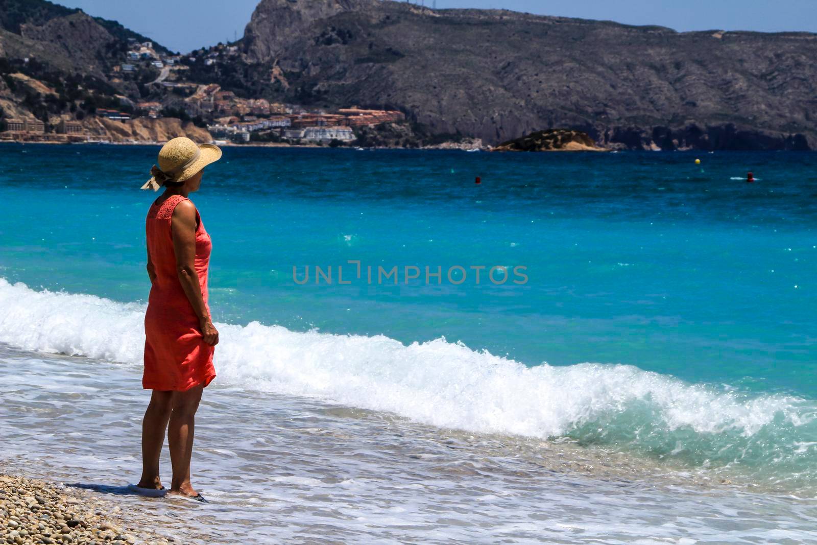 Altea, Alicante, Spain- June 12, 2021: Woman in red dress and hat on Cap Negret beach on a sunny day of Spring in Altea, Alicante, Spain