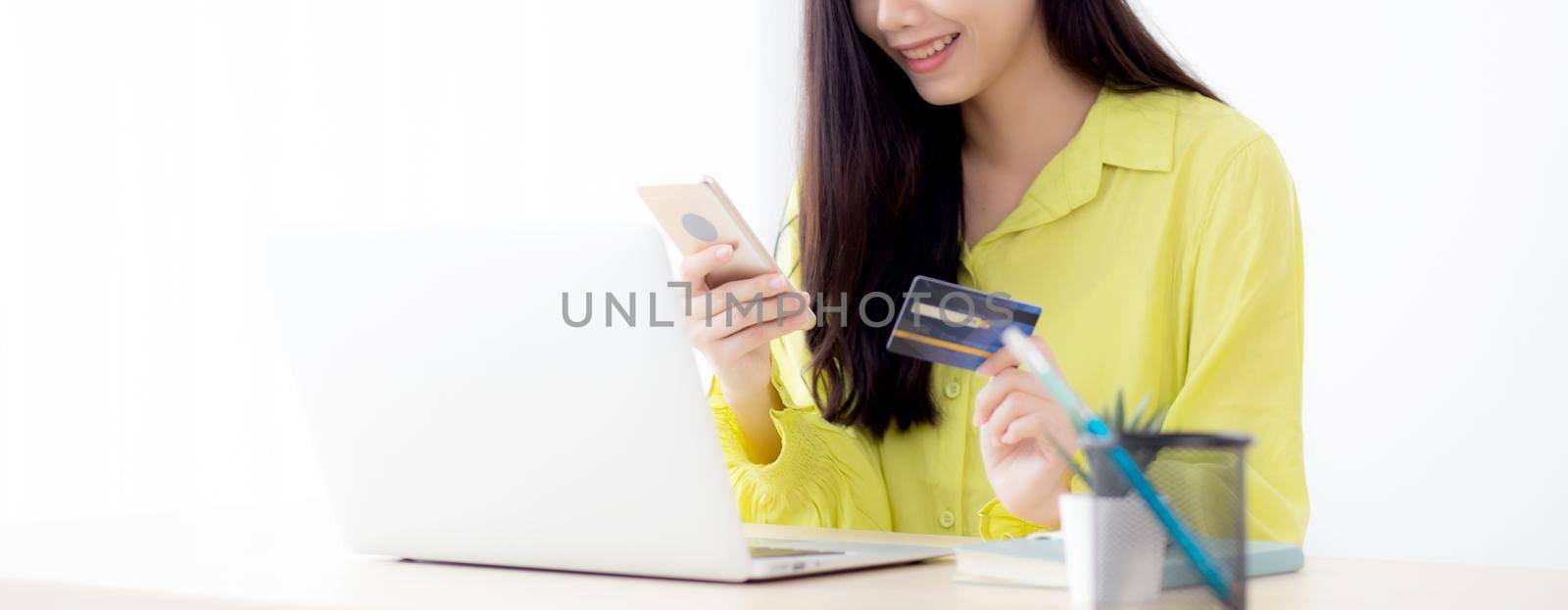 Young asian business woman using smart phone and holding credit card while online shopping and payment online with laptop computer on desk at home, female holding debit card, communication concept.