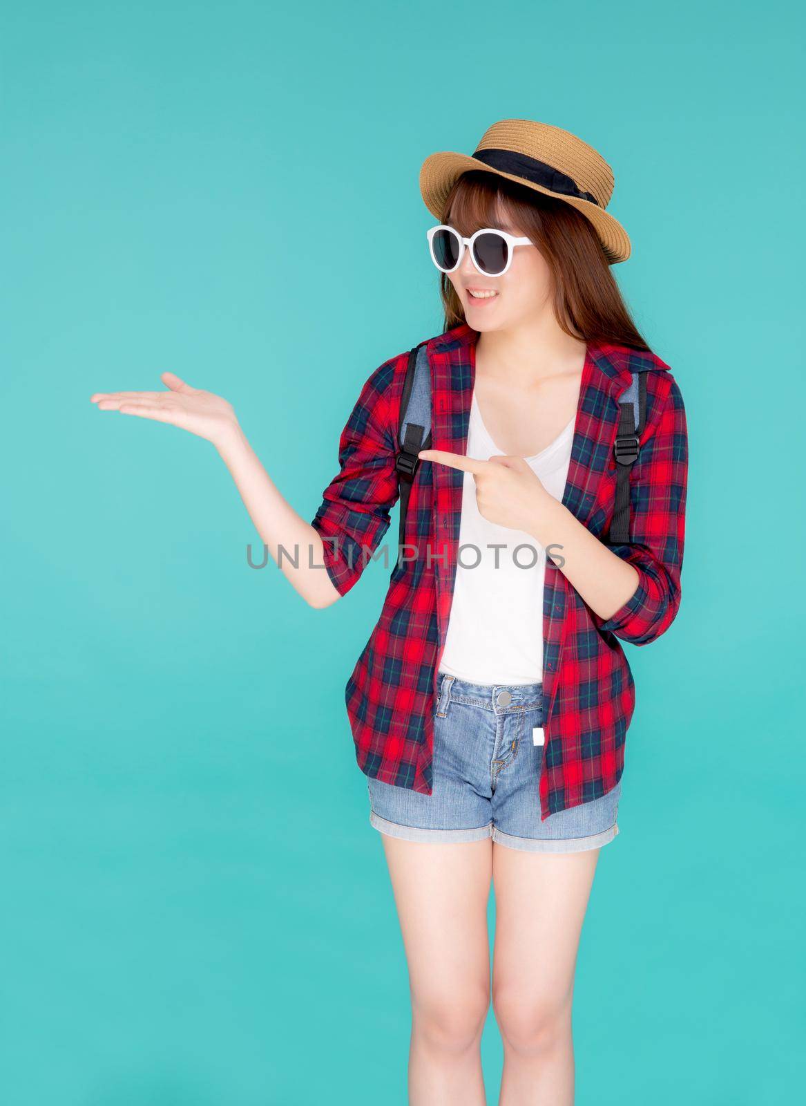 Beautiful portrait young asian woman wear sunglasses and hat smile and enjoy in summer gesture presenting and pointing something isolated on blue background, tourist girl in vacation travel concept.