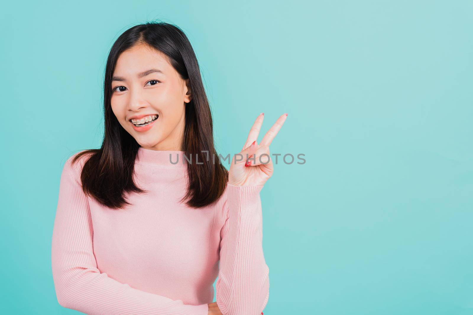 Young Asian beautiful woman smiling wear silicone orthodontic retainers on teeth showing v-sign victory finger isolated on blue background, retaining after removable braces. Dental hygiene concept