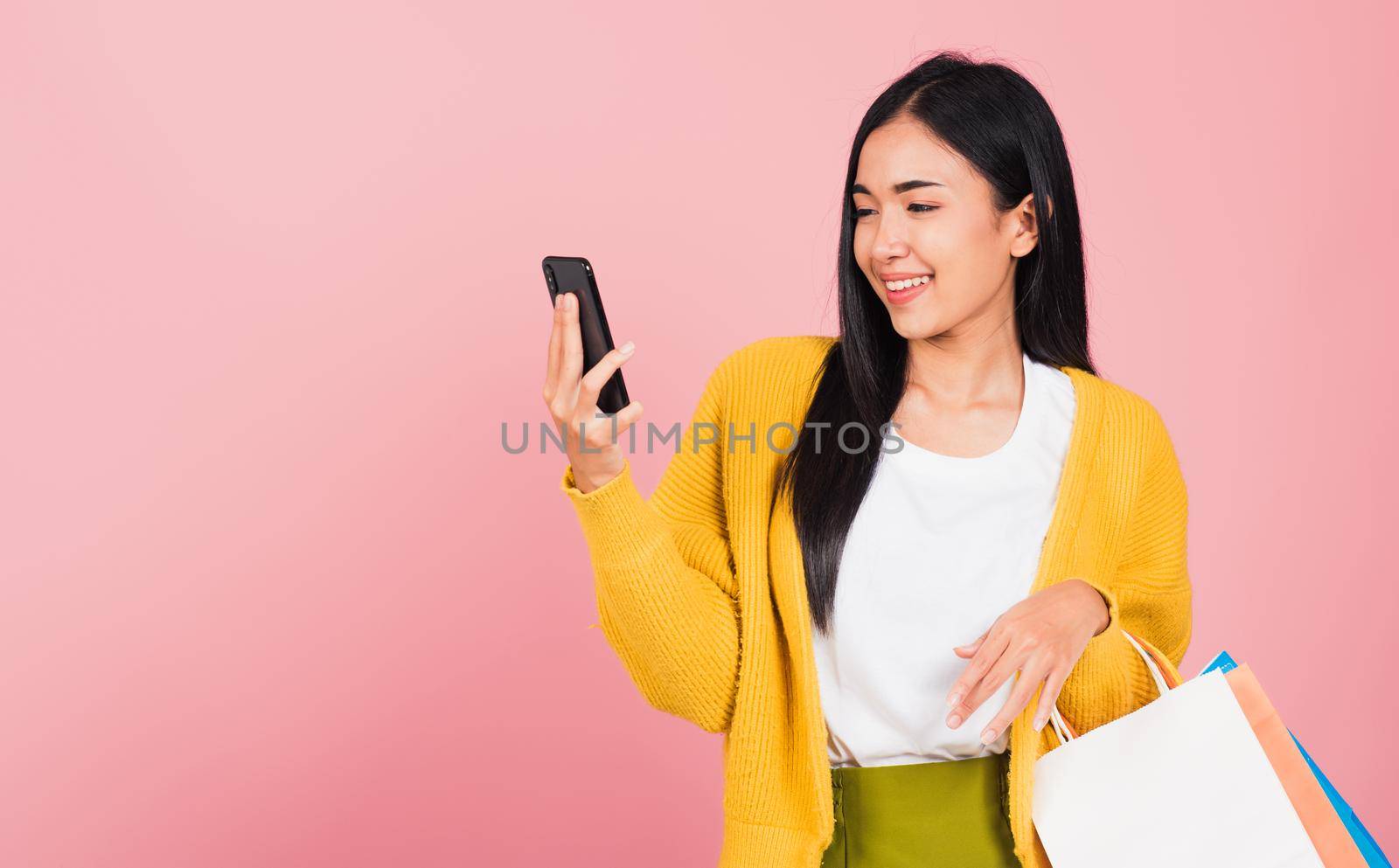 Portrait Asian happy beautiful young woman teen shopper smiling standing excited holding online shopping bags online colorful multicolor and smartphone on hand, studio shot isolated on pink background