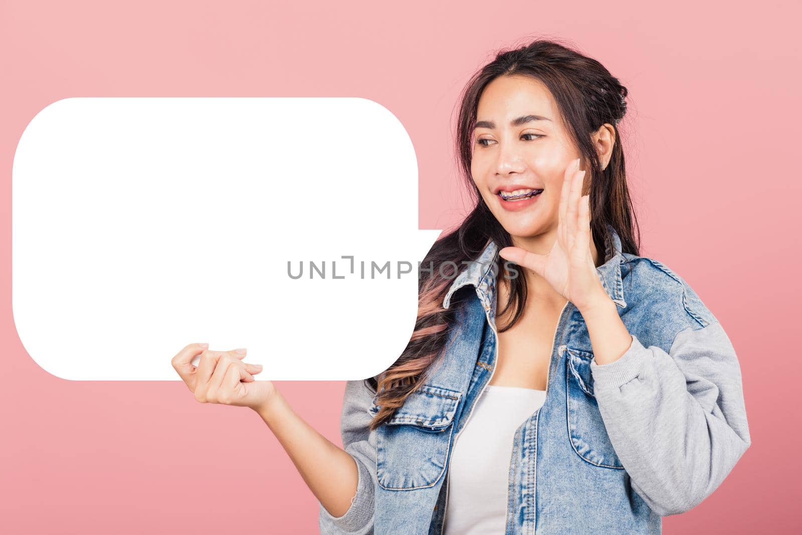Happy Asian beautiful young woman smiling excited wear denims hand near mouth announce news and holding empty speech bubble sign, Portrait female posing idea, studio shot isolated on pink background