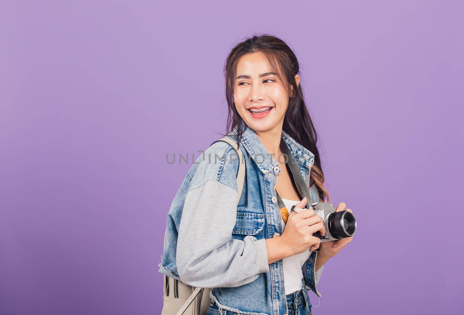 Attractive portrait happy Asian beautiful young woman smiling excited wear denims and bag holding vintage photo camera, female traveler female photographer, studio shot isolated on purple background