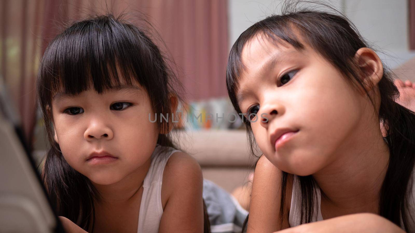 Two sibling girls lying on the sofa and watching digital tablets together in the living room. by TEERASAK