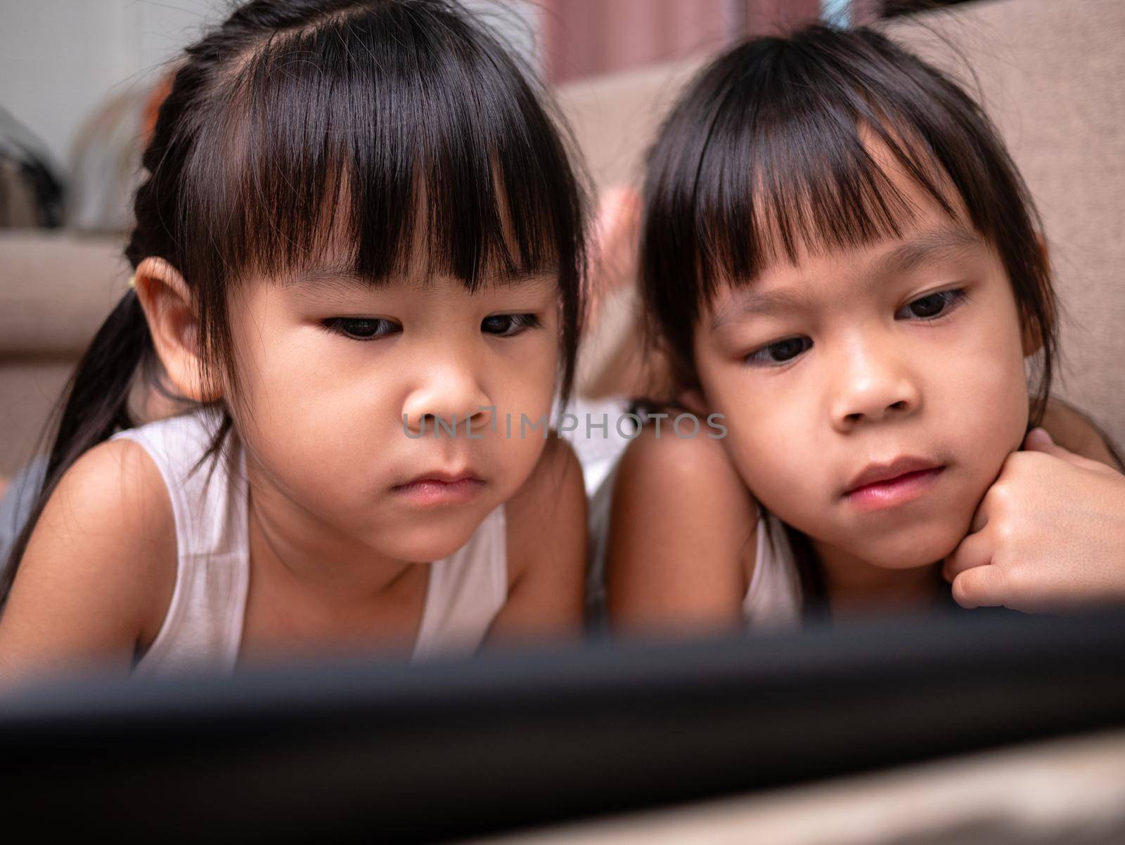 Two sibling girls lying on the sofa and watching digital tablets together in the living room.