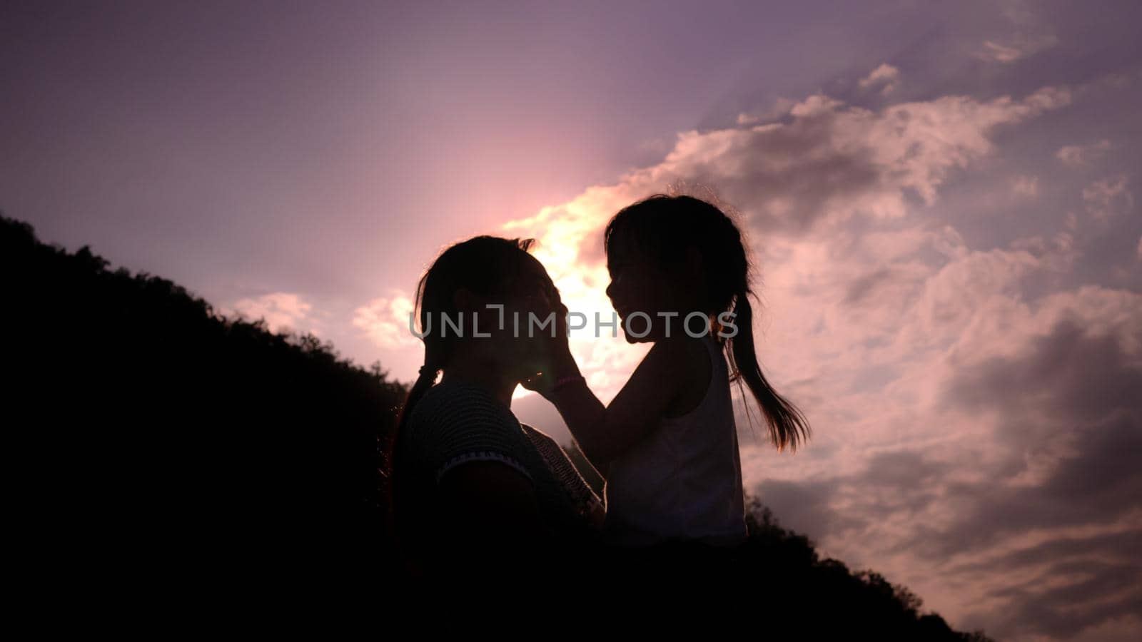 The silhouette of happy mother holding little girl smiling and playing on sunset background in the park. by TEERASAK