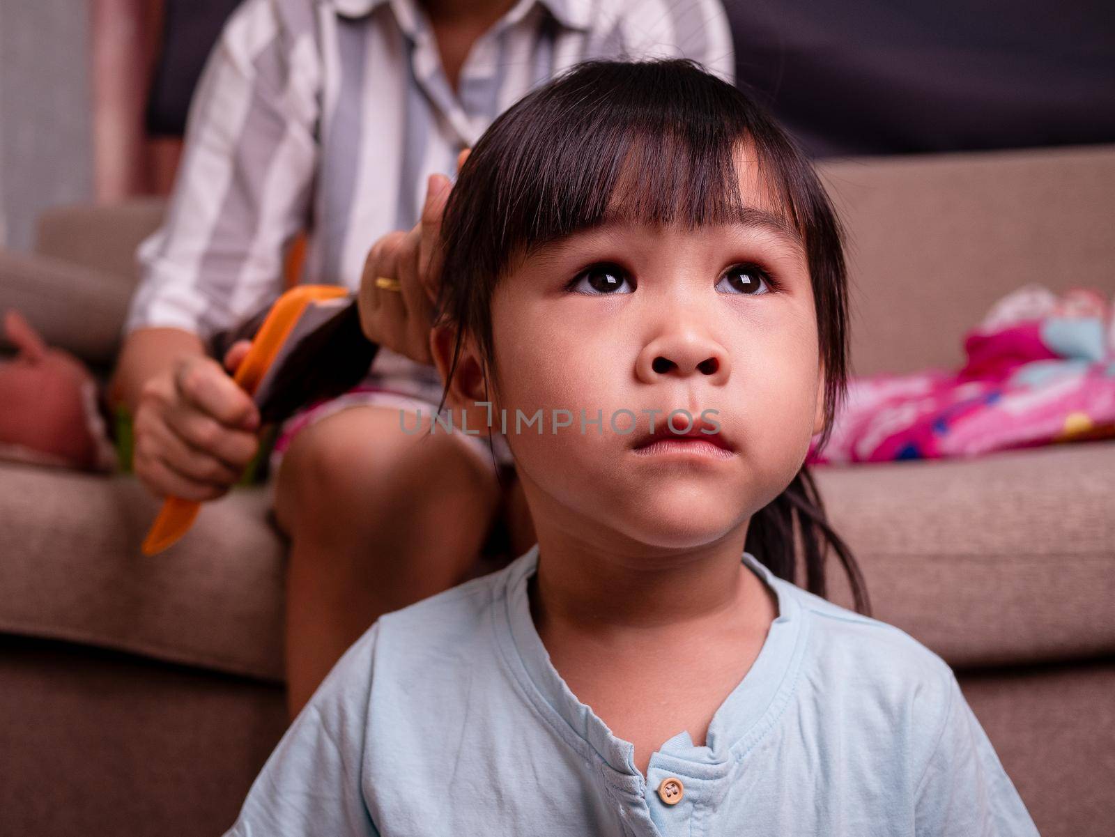 Close up of charming little girl is looking at TV while her mother is combing daughter's hair in living room.