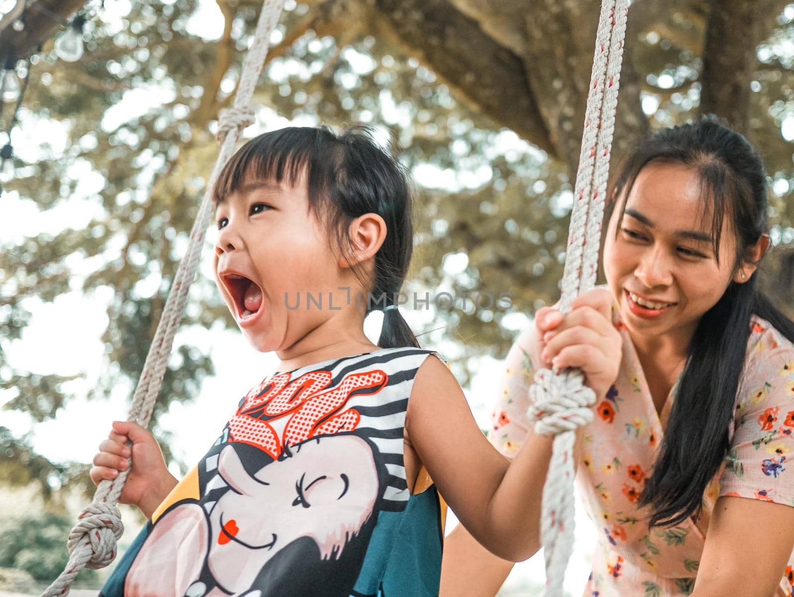 Happy mother pushing laughing daughter on swing in summer park. Family spend time together on vacation.