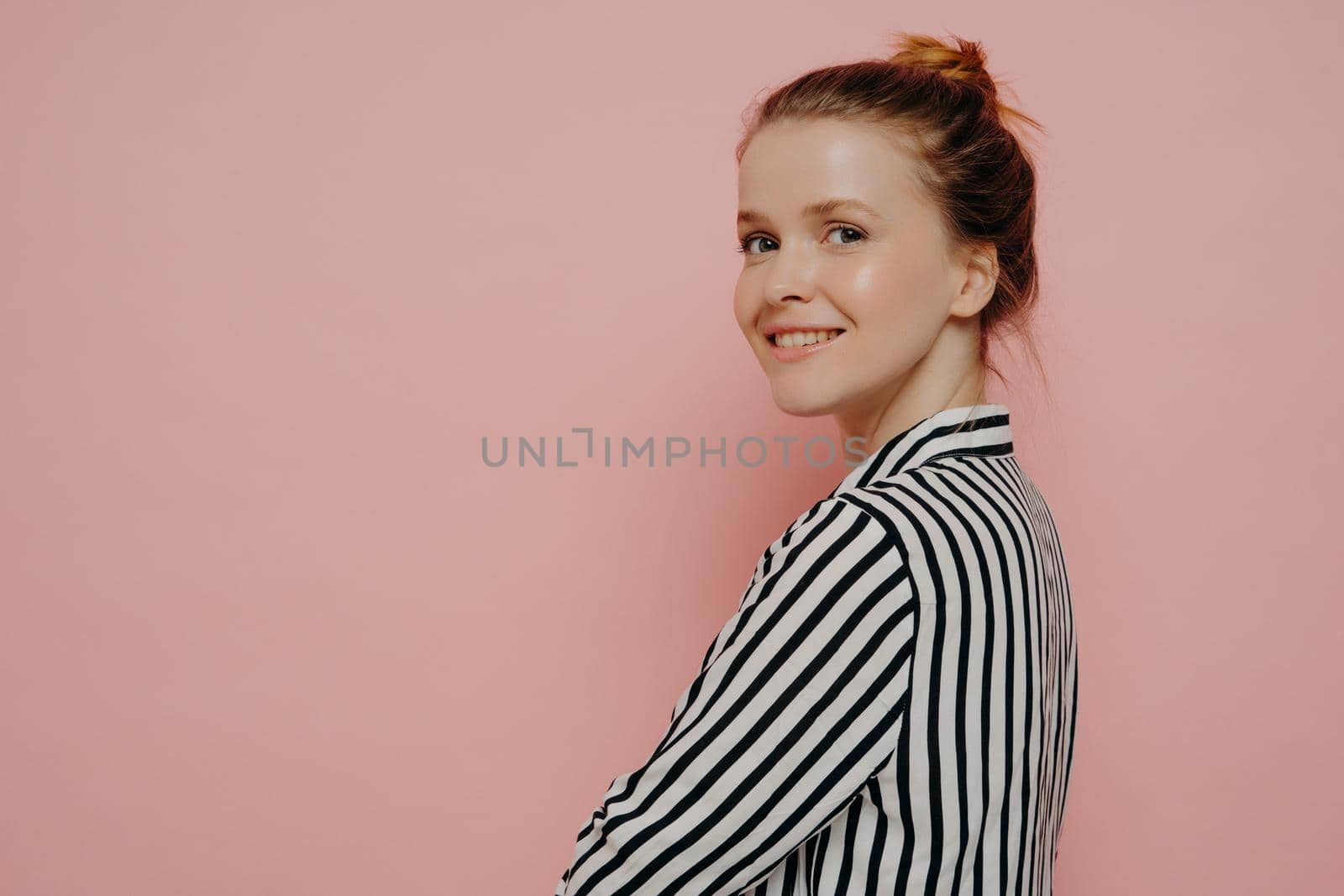 Relaxed brunette teenage girl dressed in white and black shirt with hair in bun, looking at camera with crossed arms while smiling, feeling happy while standing alone in front of pink background