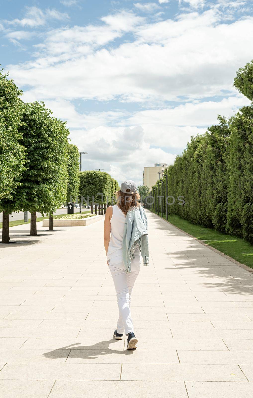 Woman in casual jeans clothes walking in the park. Back view of a blond woman holding her cap walking outdoors