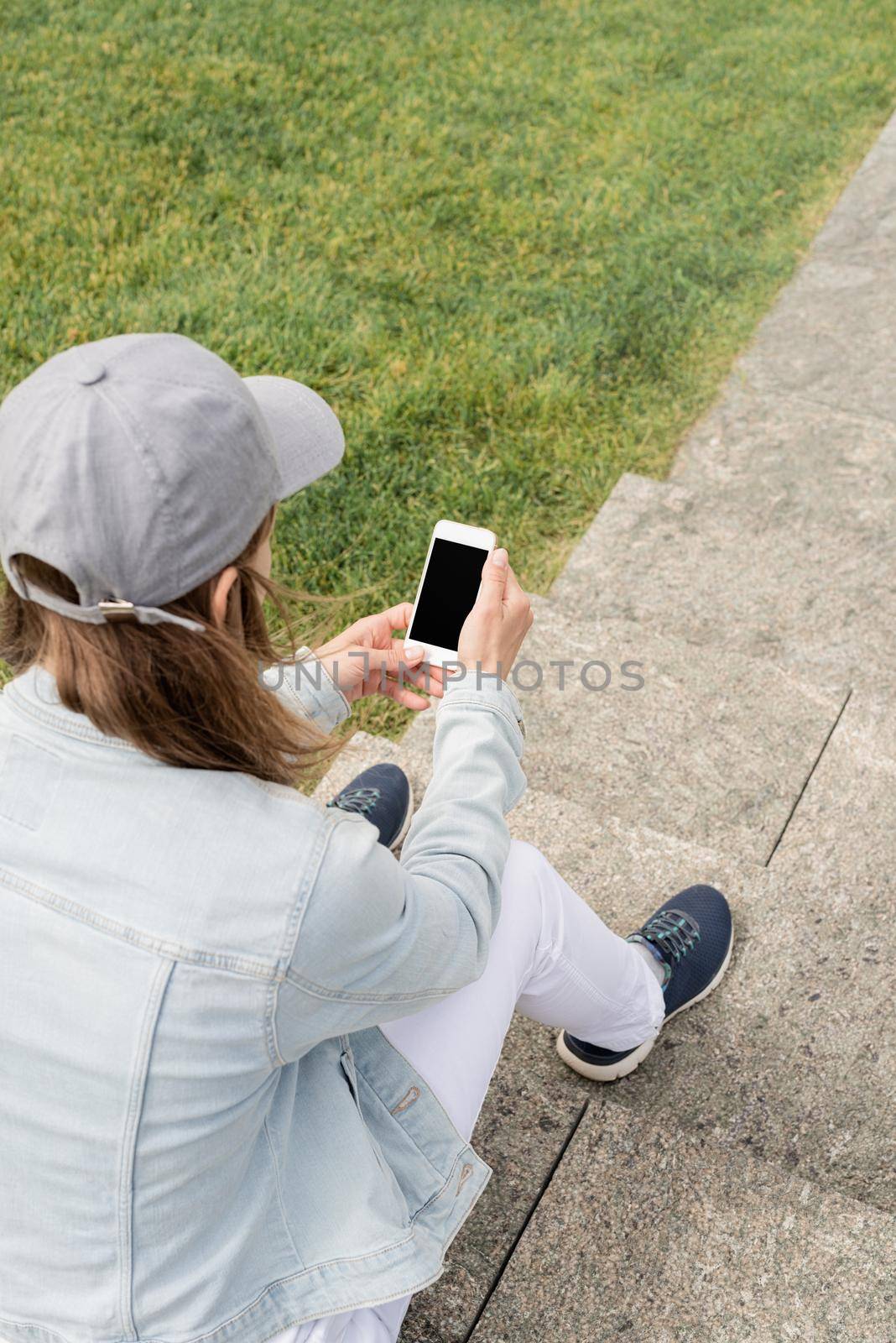 Woman in jeans clothes texting on her mobile phone, sitting on the stairs in the park. Mock up design