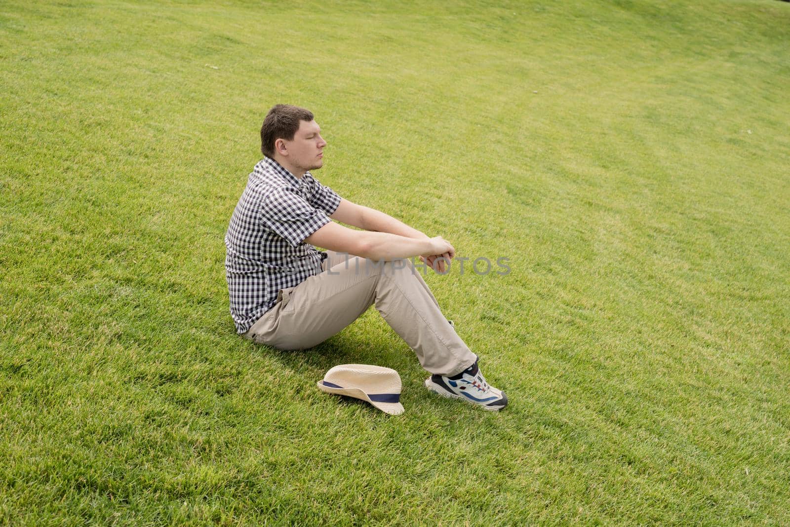 Young thoughtful man in summer hat sitting on the grass in the park, looking away