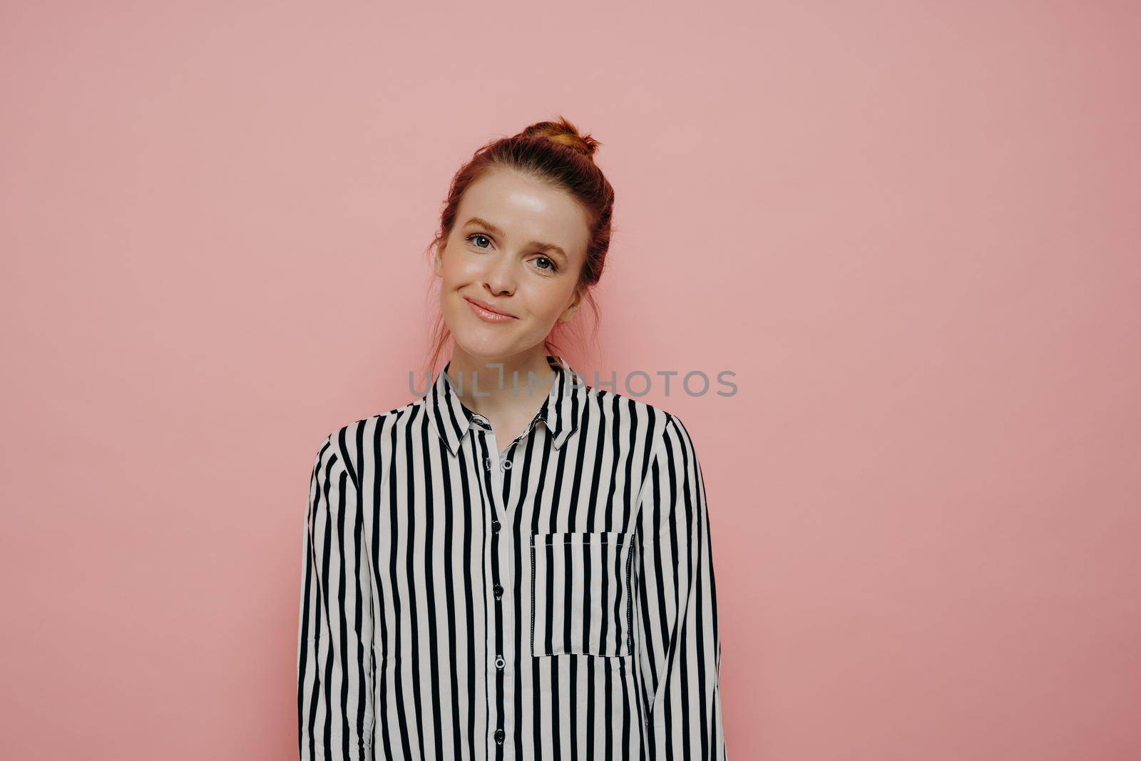 Portrait of beautiful glad young woman looking at camera with slight smile and tilted head, admiring something while standing isolated over pink background, attractive young smiling female student