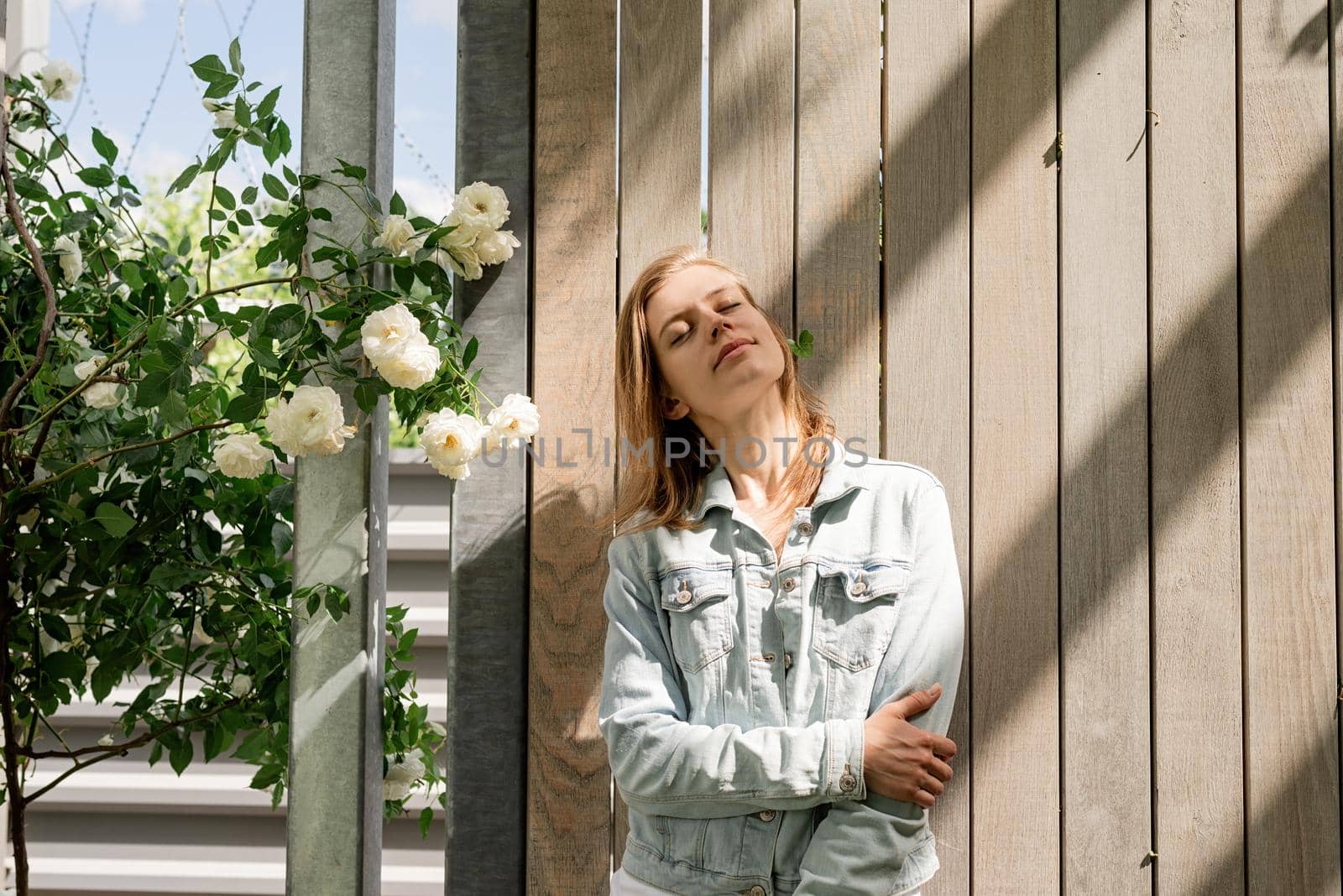 young woman standing in white roses in a garden or park, wooden background by Desperada