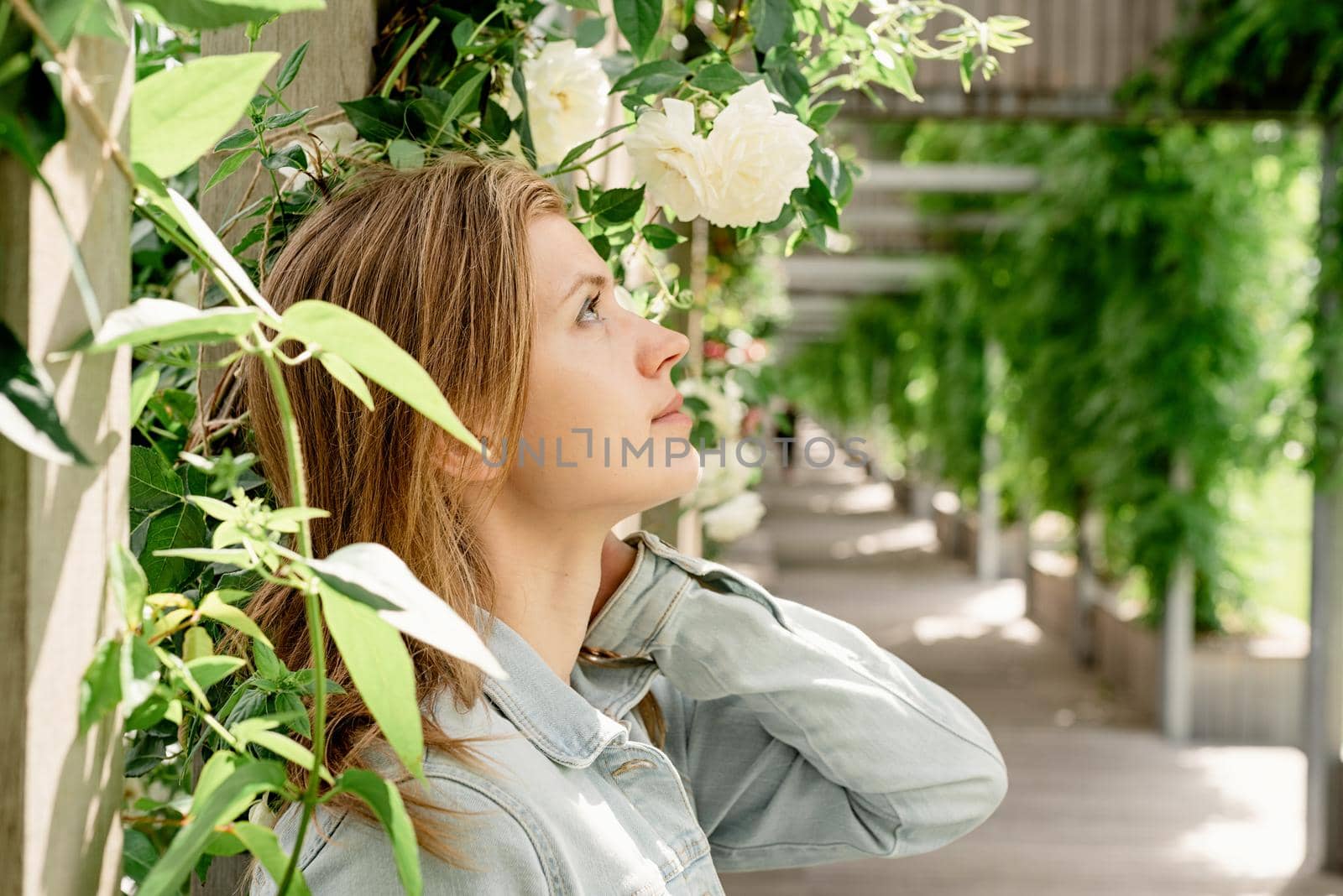 Beautiful young blond woman standing in white roses in a garden or park looking away
