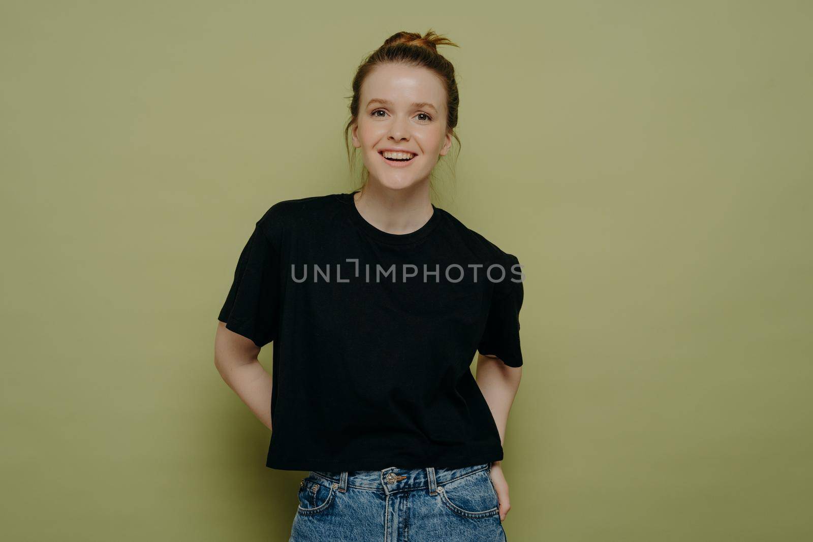 Young happy young woman with hair in bun dressed in casual clothes standing in relaxed posture and looking at camera with wide smile on face while posing in studio on green background